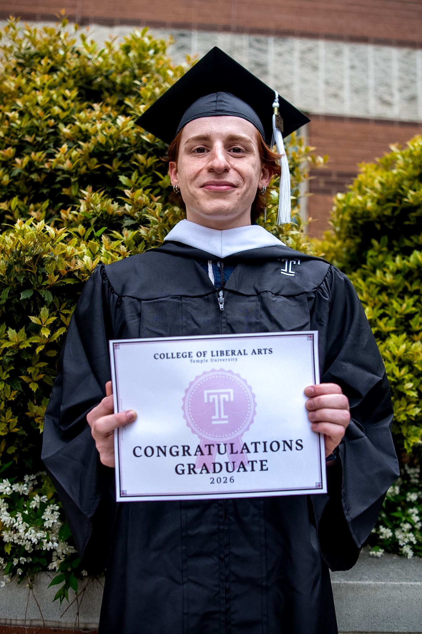 Temple University College of Liberal Arts 2026 graduate in cap and gown holding congratulations sign outdoors.