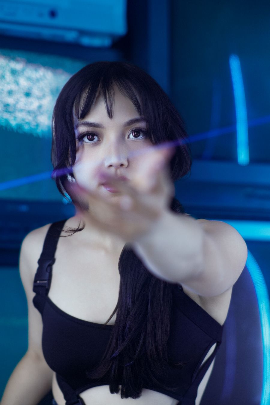 Young woman in black crop top reaches toward camera with purple light streak in blue-lit studio.