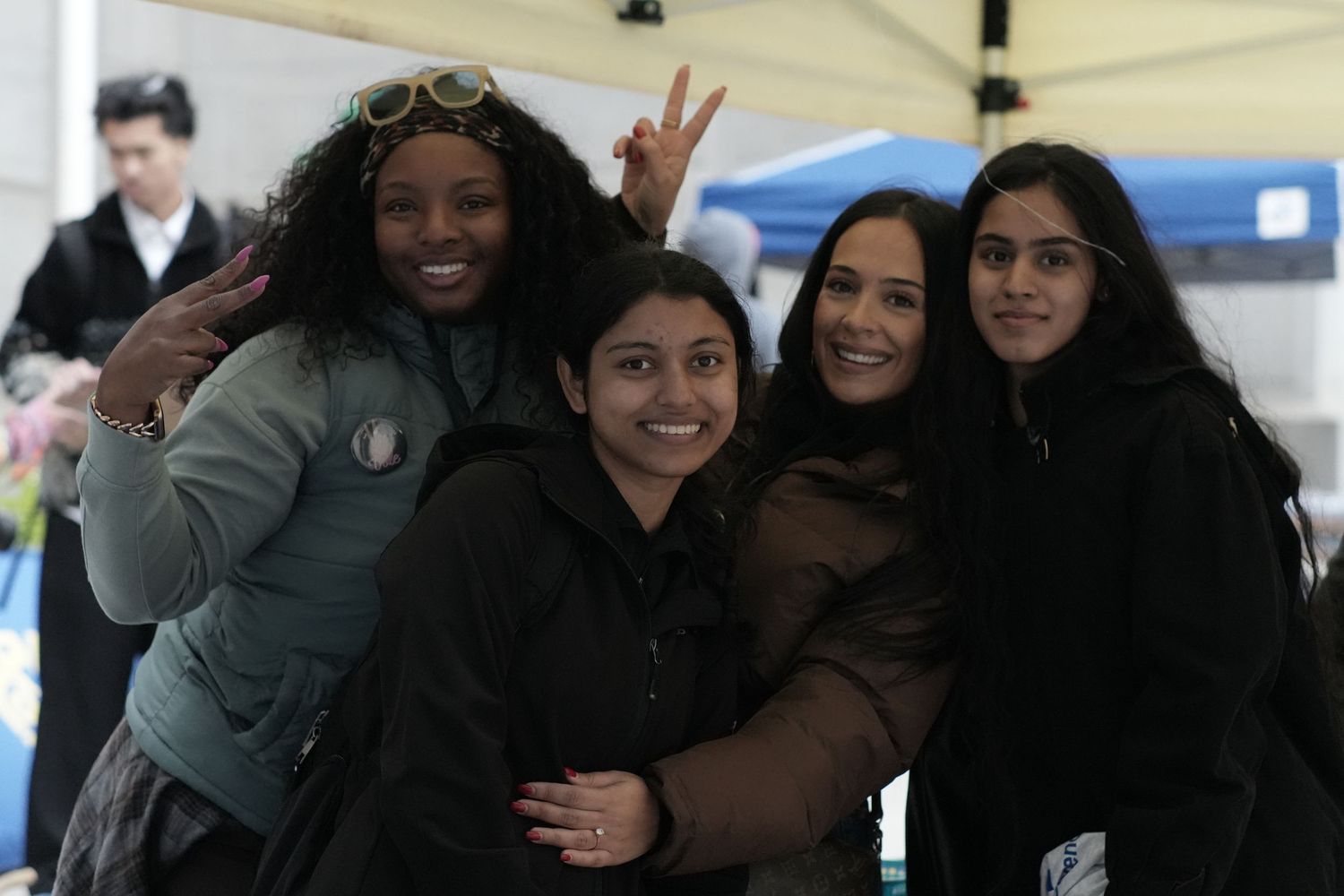 Four smiling young women posing together outdoors under a tent at a community event.