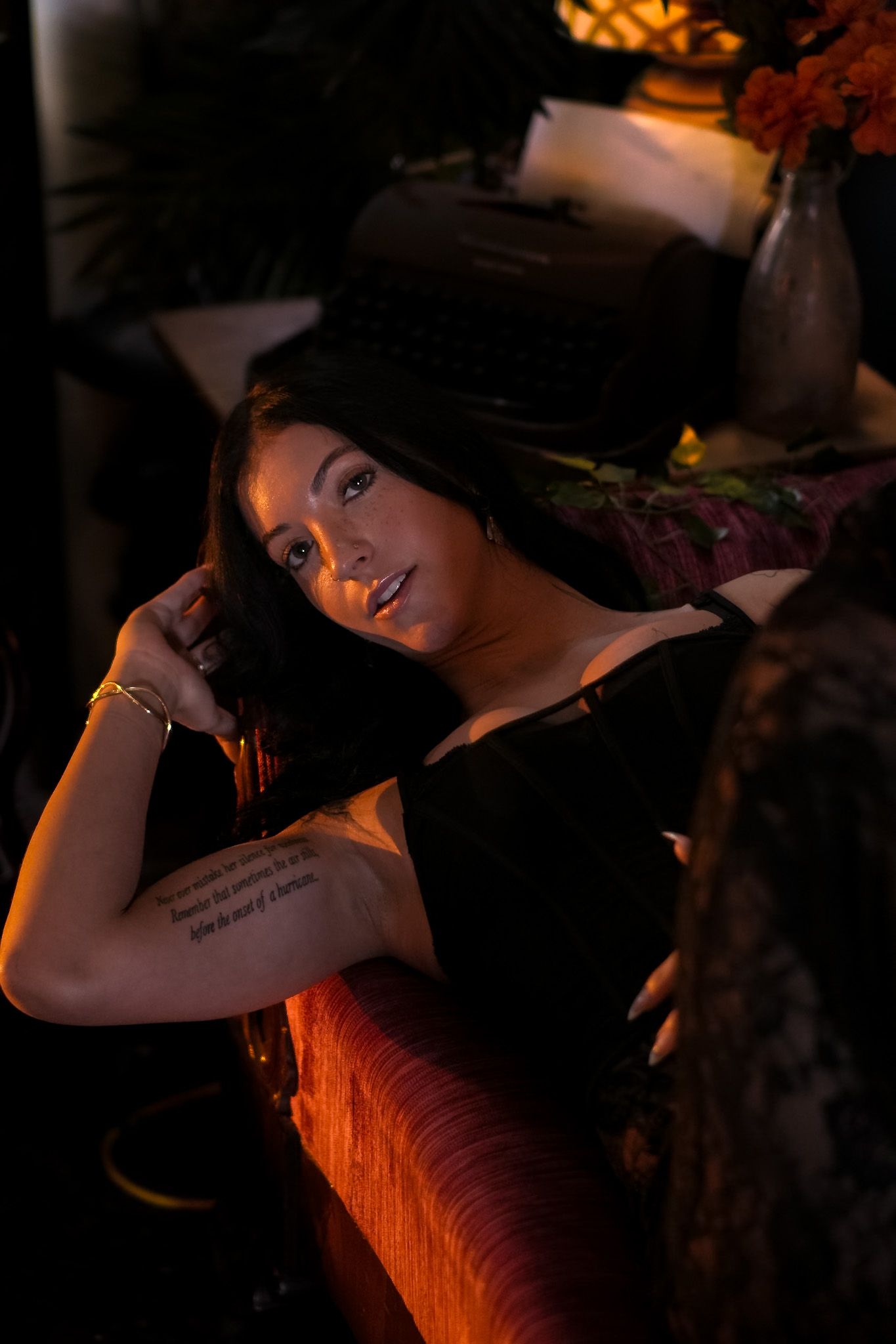 Brunette woman with arm tattoo relaxing on a couch in warm ambient lighting at an intimate venue.