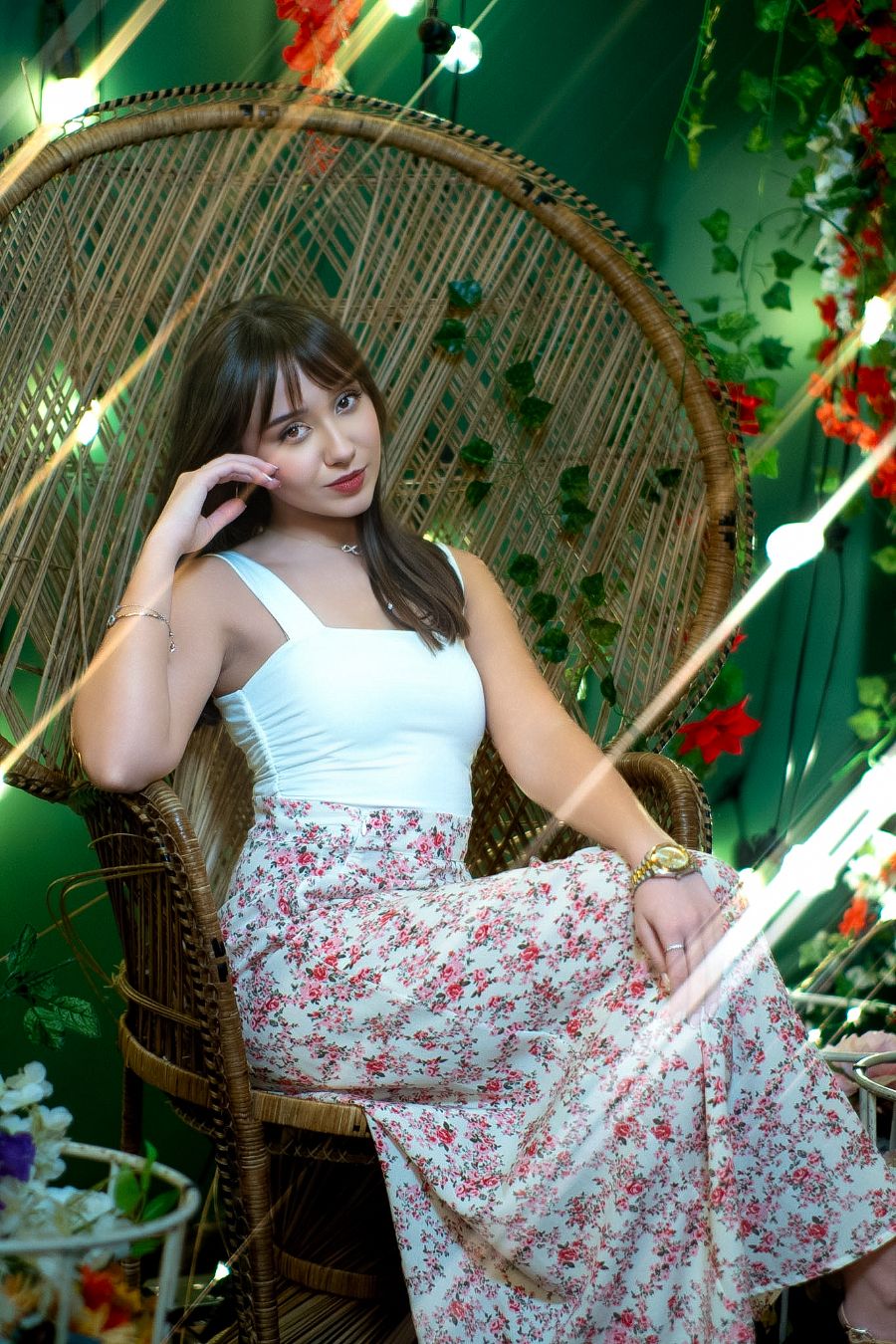 Young woman in white top and floral skirt sitting in wicker peacock chair surrounded by colorful flowers.