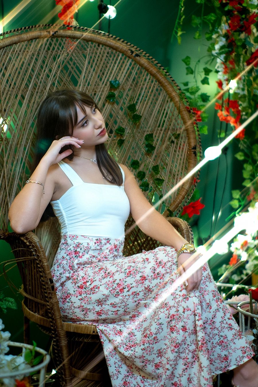 Young woman in white top and floral skirt sitting in a wicker peacock chair surrounded by green vines and flowers.