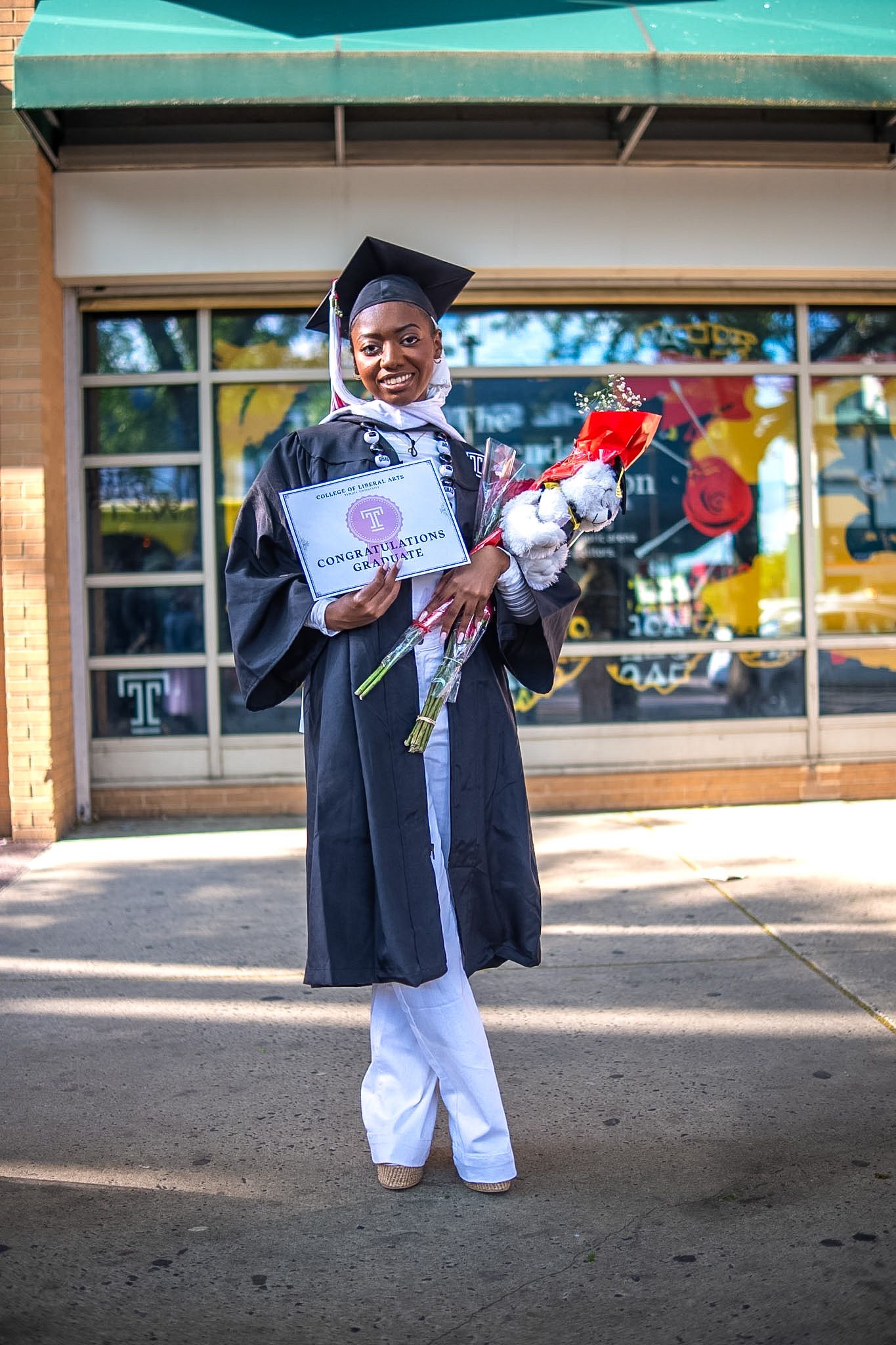Smiling graduate in black cap and gown holds diploma and flowers outside on a sunny day.