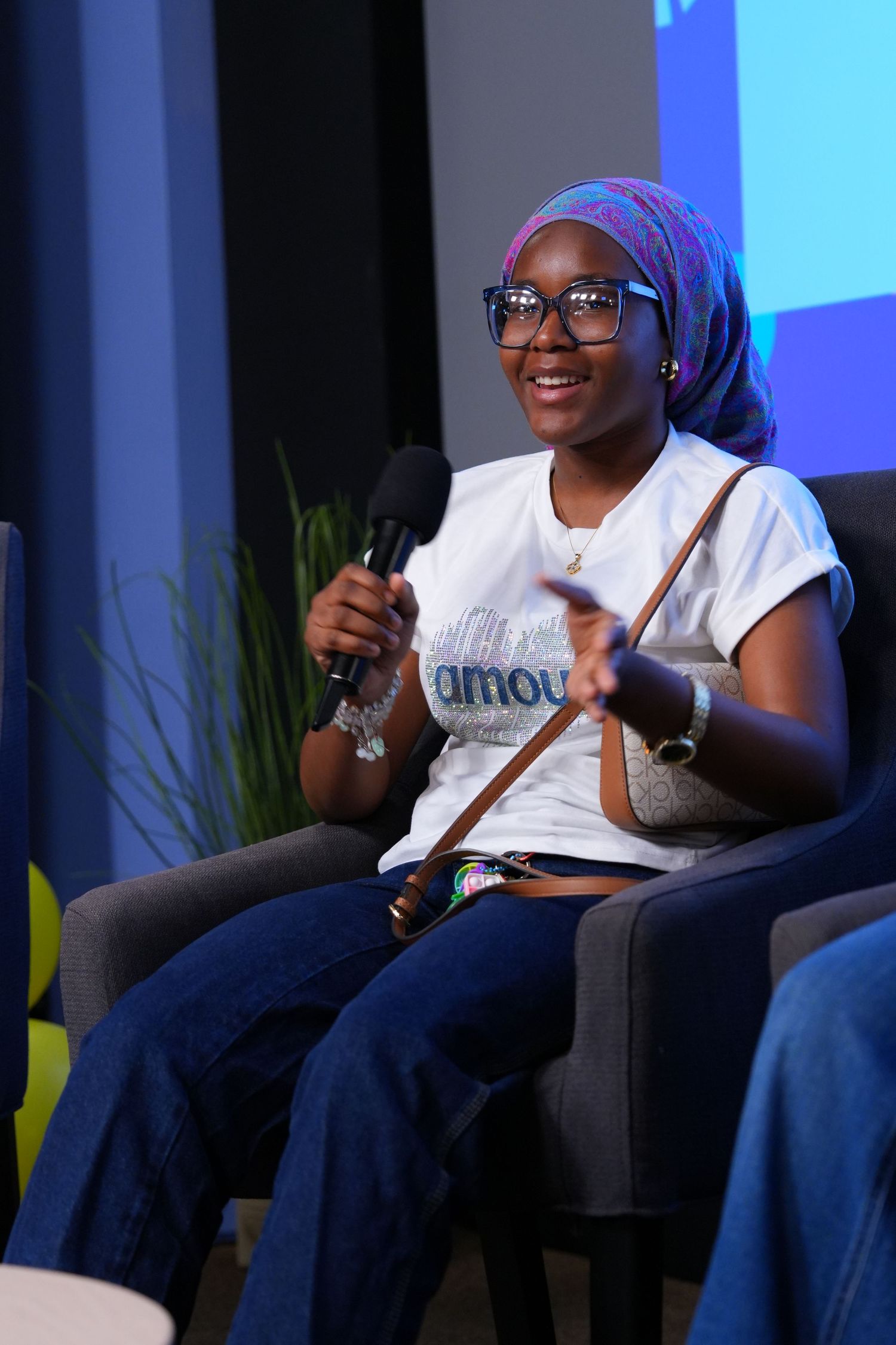 Young woman in hijab and glasses speaks into microphone while seated at a panel discussion event.