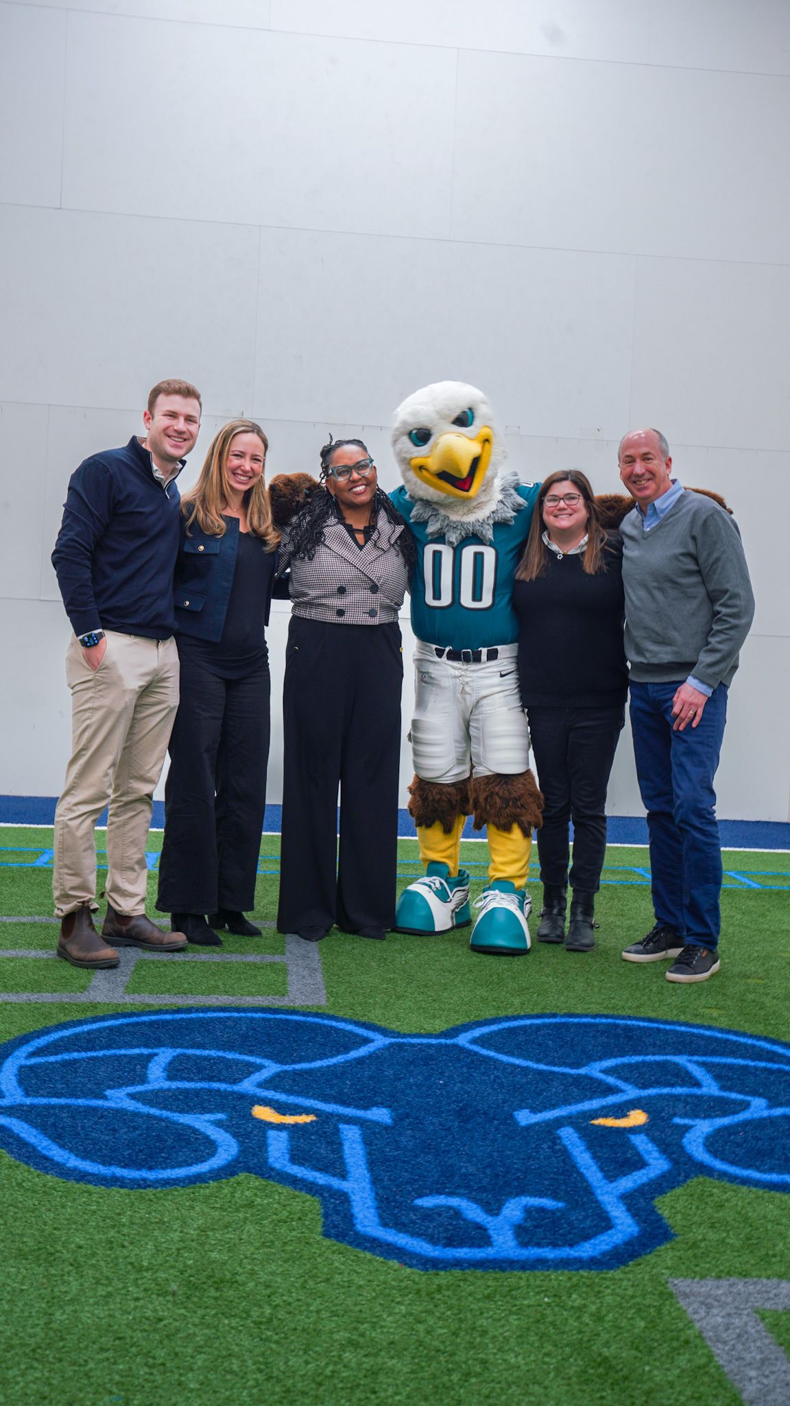 Group of five adults posing with Philadelphia Eagles mascot Swoop on an indoor turf field.