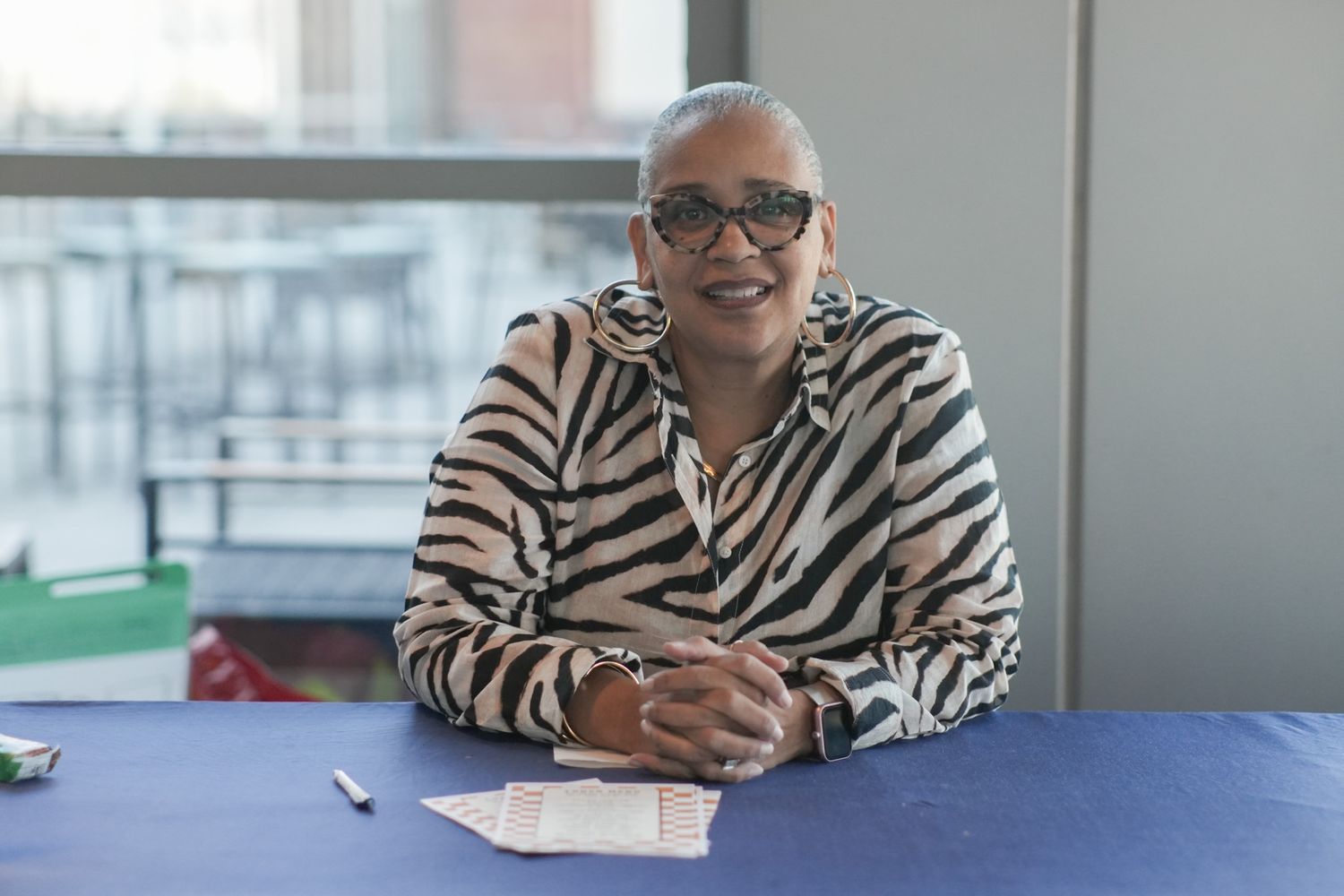 Smiling older woman in zebra-print blazer sitting at a blue table with papers and a pen.