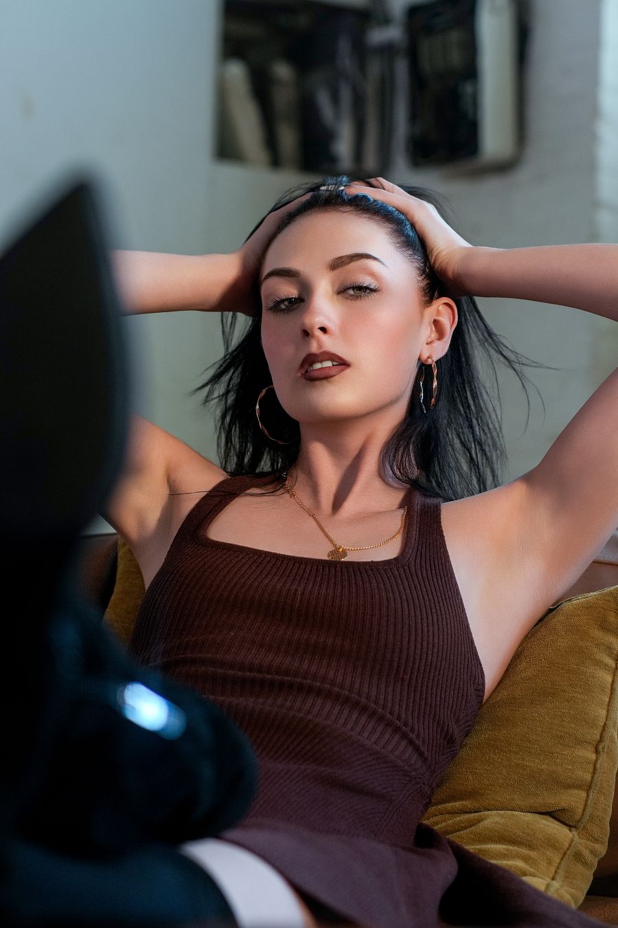 Young woman with dark hair in brown tank top posing with hands behind head on gold velvet sofa.