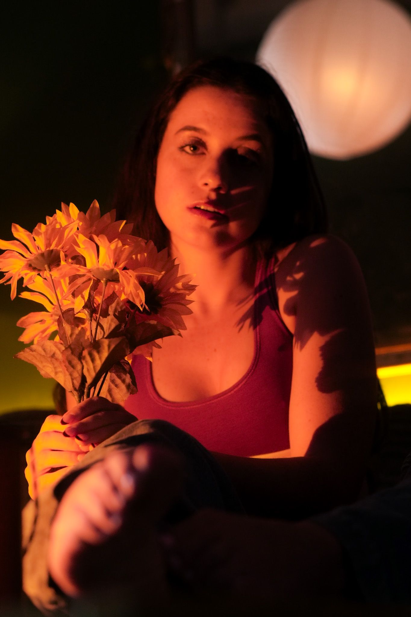 Young brunette woman holding sunflowers in dramatic warm orange lighting against dark background.