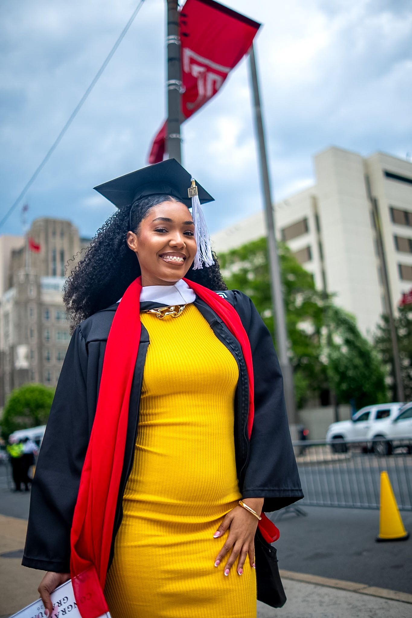 Smiling woman in graduation cap, gown, and yellow dress celebrates outdoors near a red university banner.