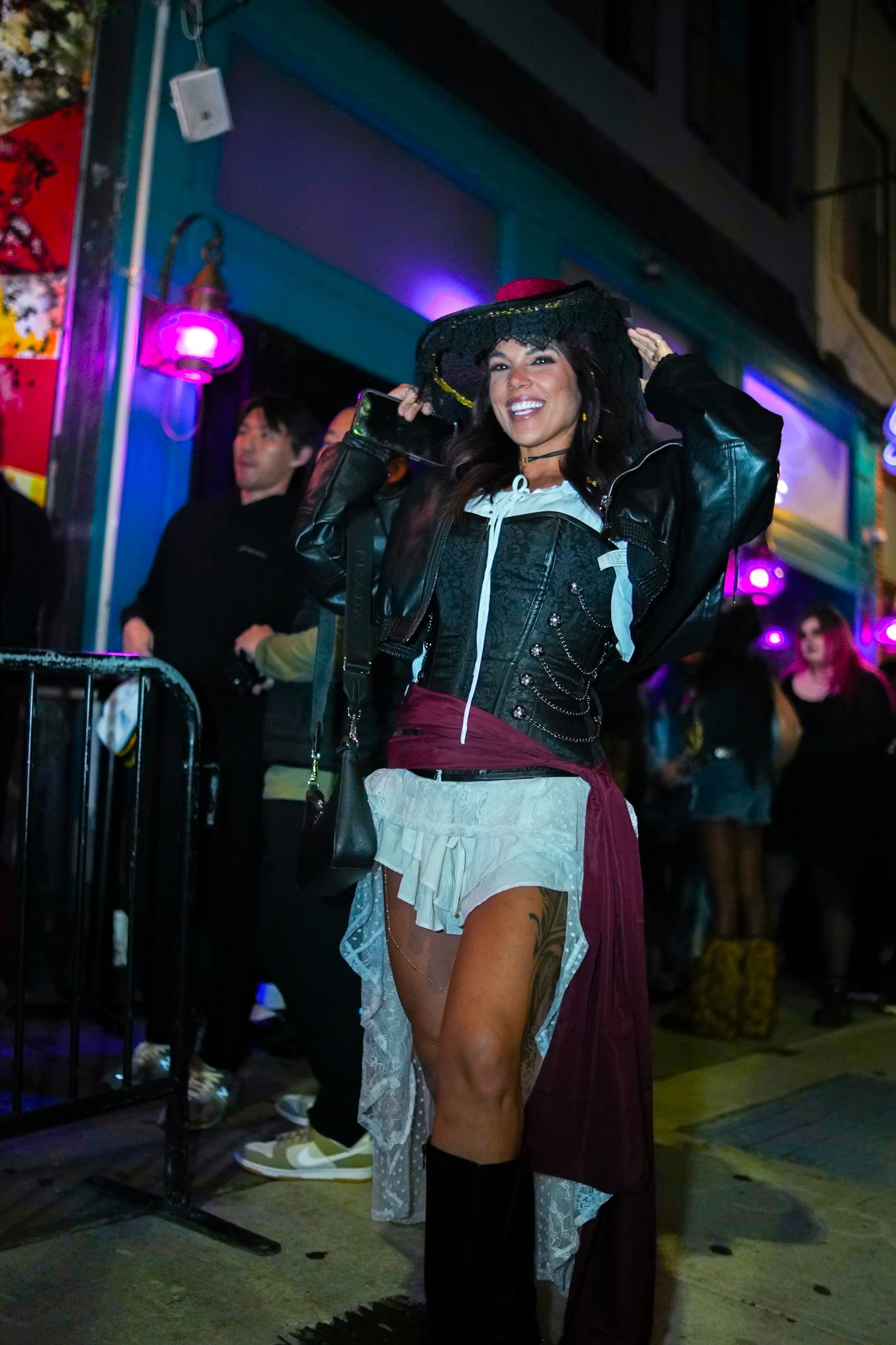 Woman in pirate Halloween costume with leather jacket and hat poses on colorful neon-lit street.