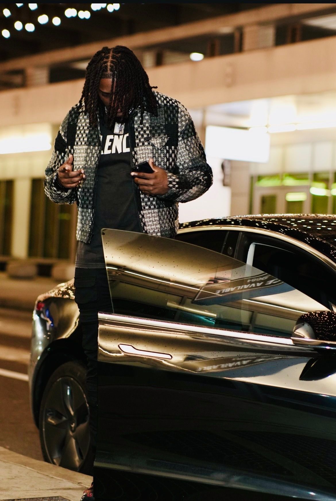 Man with braids in patterned jacket uses phone while standing beside sleek black sports car at night.