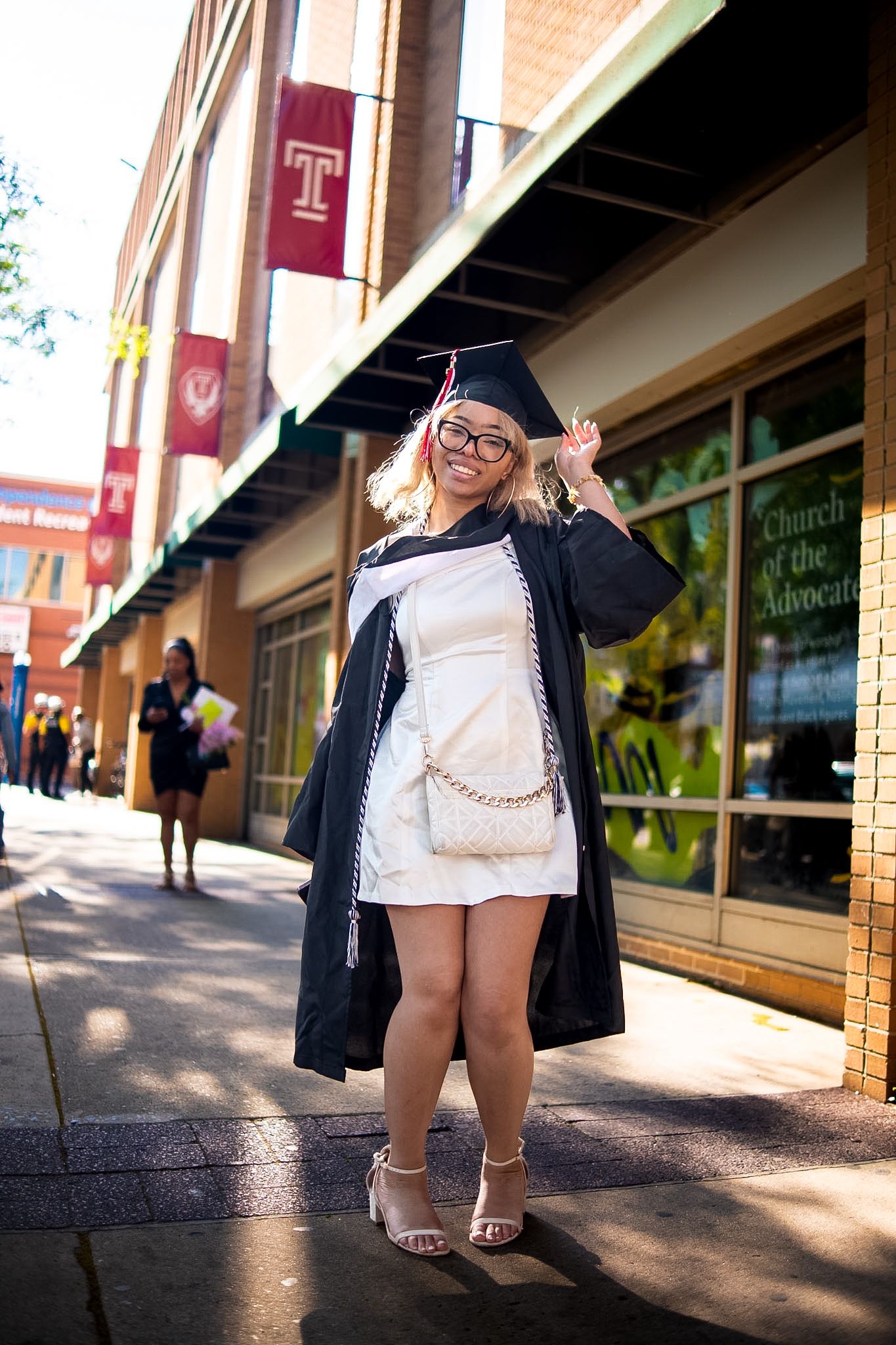 A smiling graduate in black cap and gown poses on a Temple University sidewalk wearing a white dress and heels.