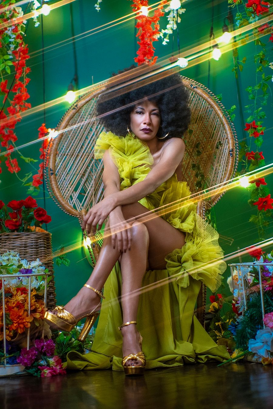 Woman in yellow ruffled gown and gold heels seated in wicker chair surrounded by colorful flowers and string lights.