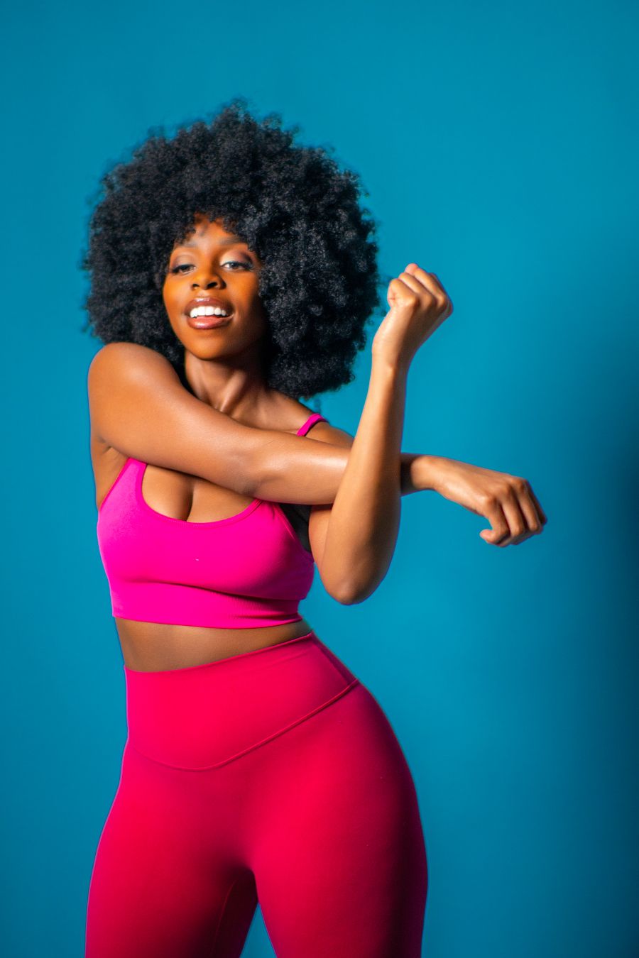 A smiling Black woman with an afro in a pink sports bra and leggings stretches her arm against a blue background.