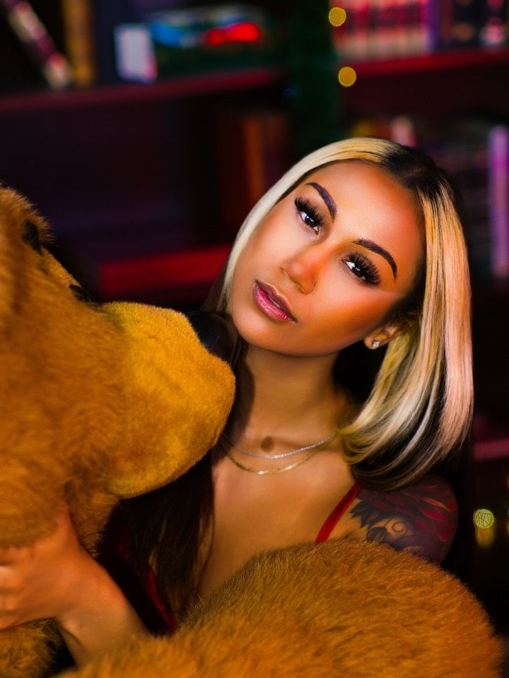 Young woman with blonde highlighted hair posing with large brown teddy bear in warm holiday lighting.