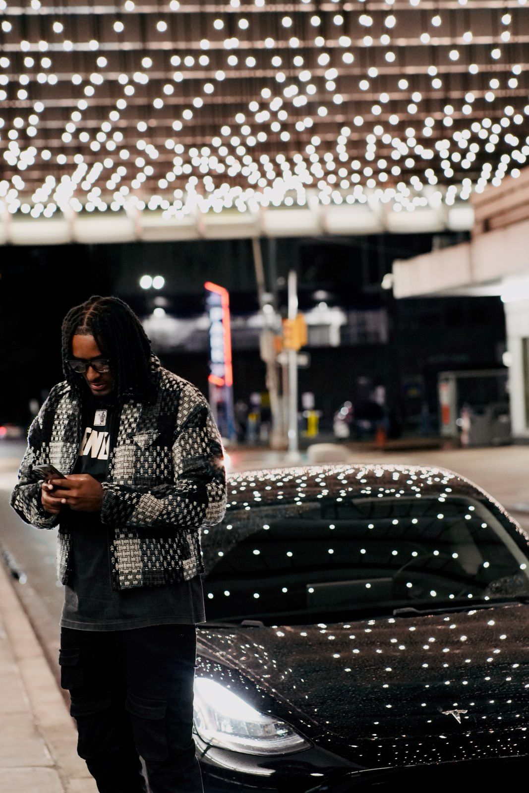 Man with braids in patterned jacket checks phone leaning on black Tesla under string lights at night.
