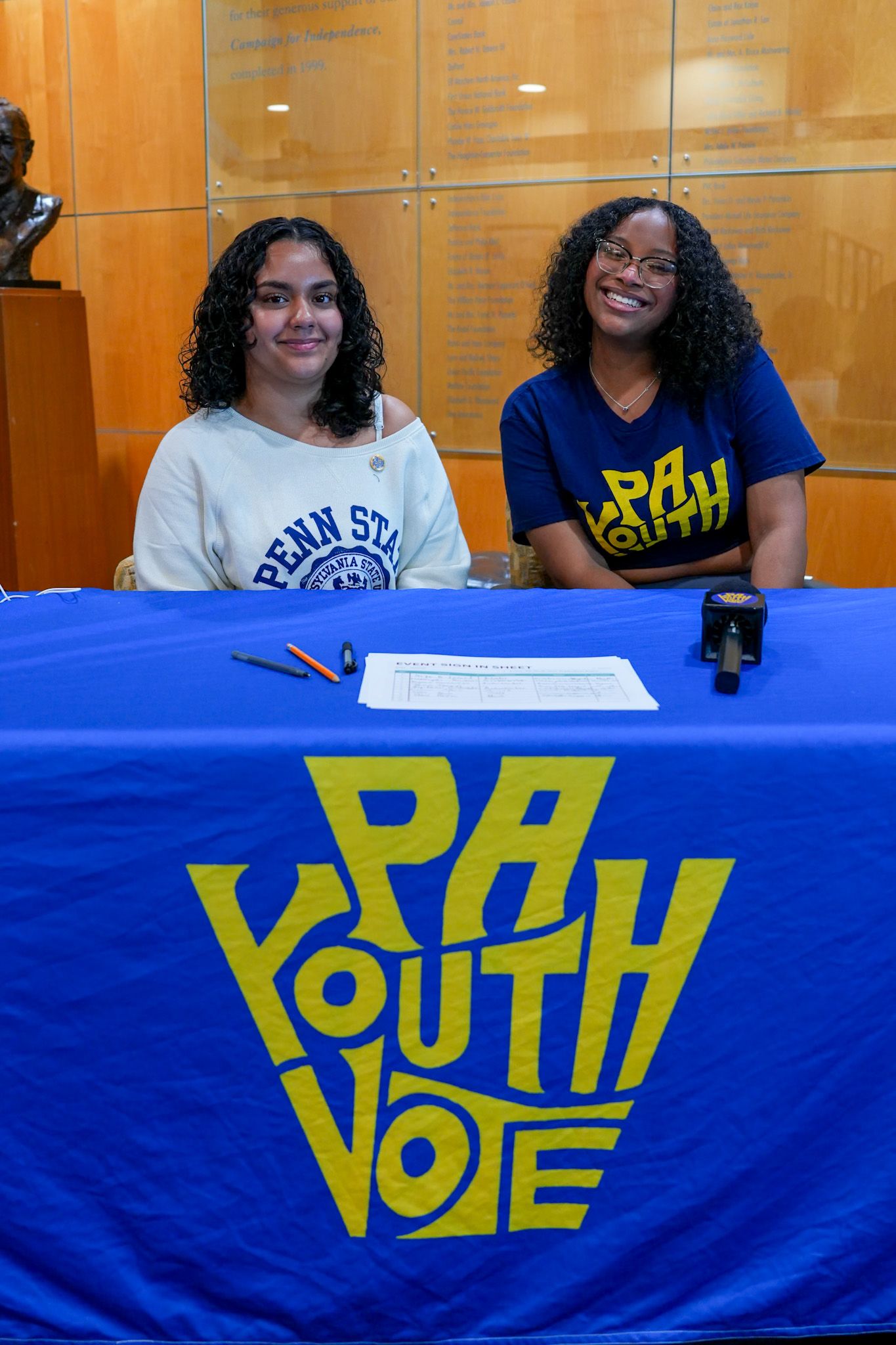 Two young women sit at a blue PA Youth Vote table, smiling, one wearing a Penn State shirt.