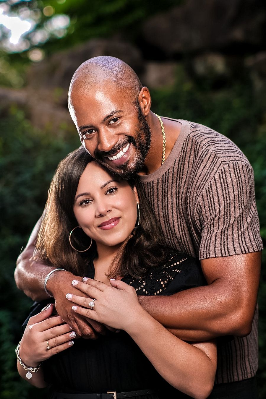 A smiling couple embraces outdoors, man with beard hugs woman wearing hoop earrings and black top.