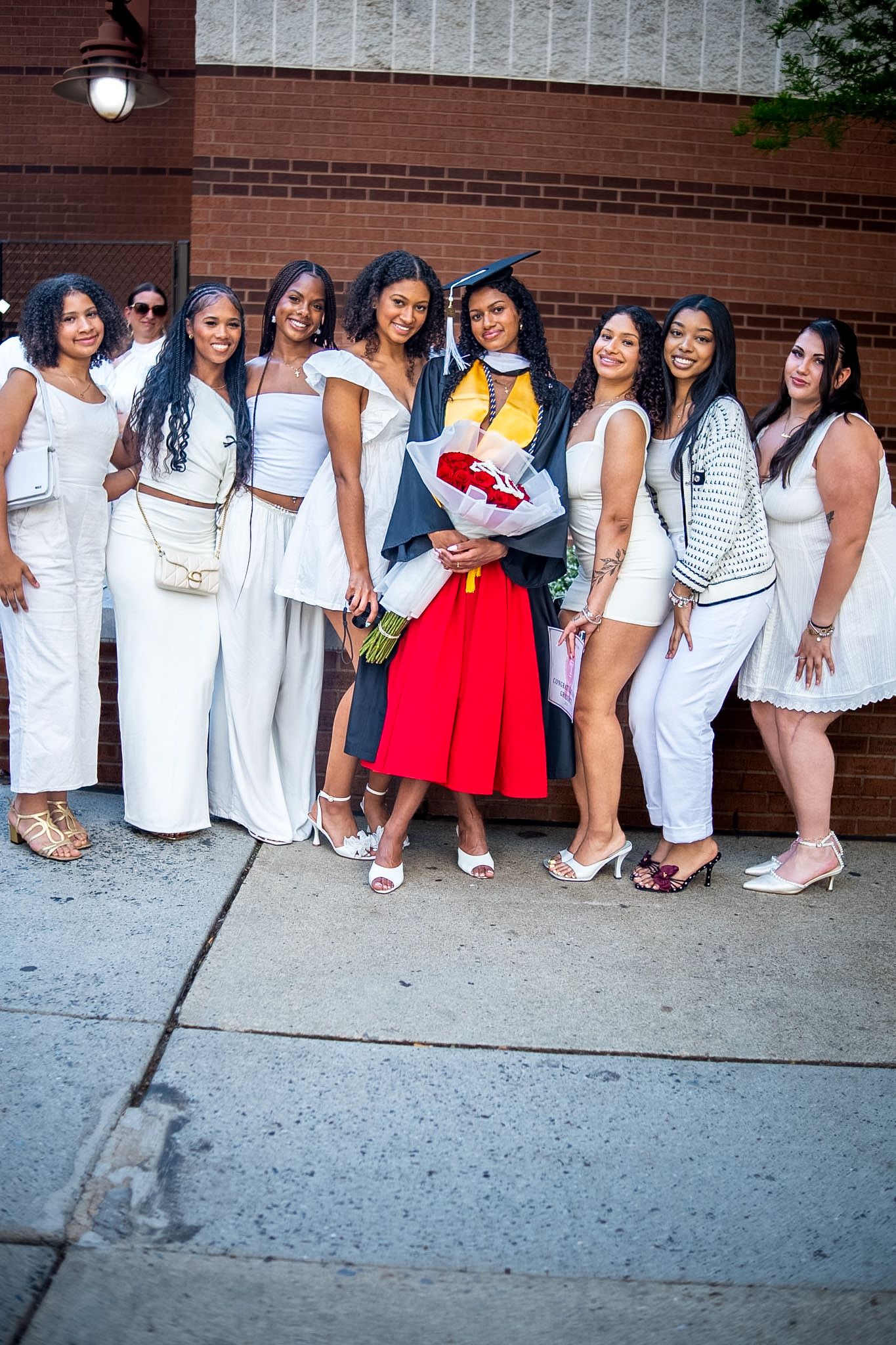 Group of women in white outfits celebrating a graduation with a graduate in cap and gown holding flowers.