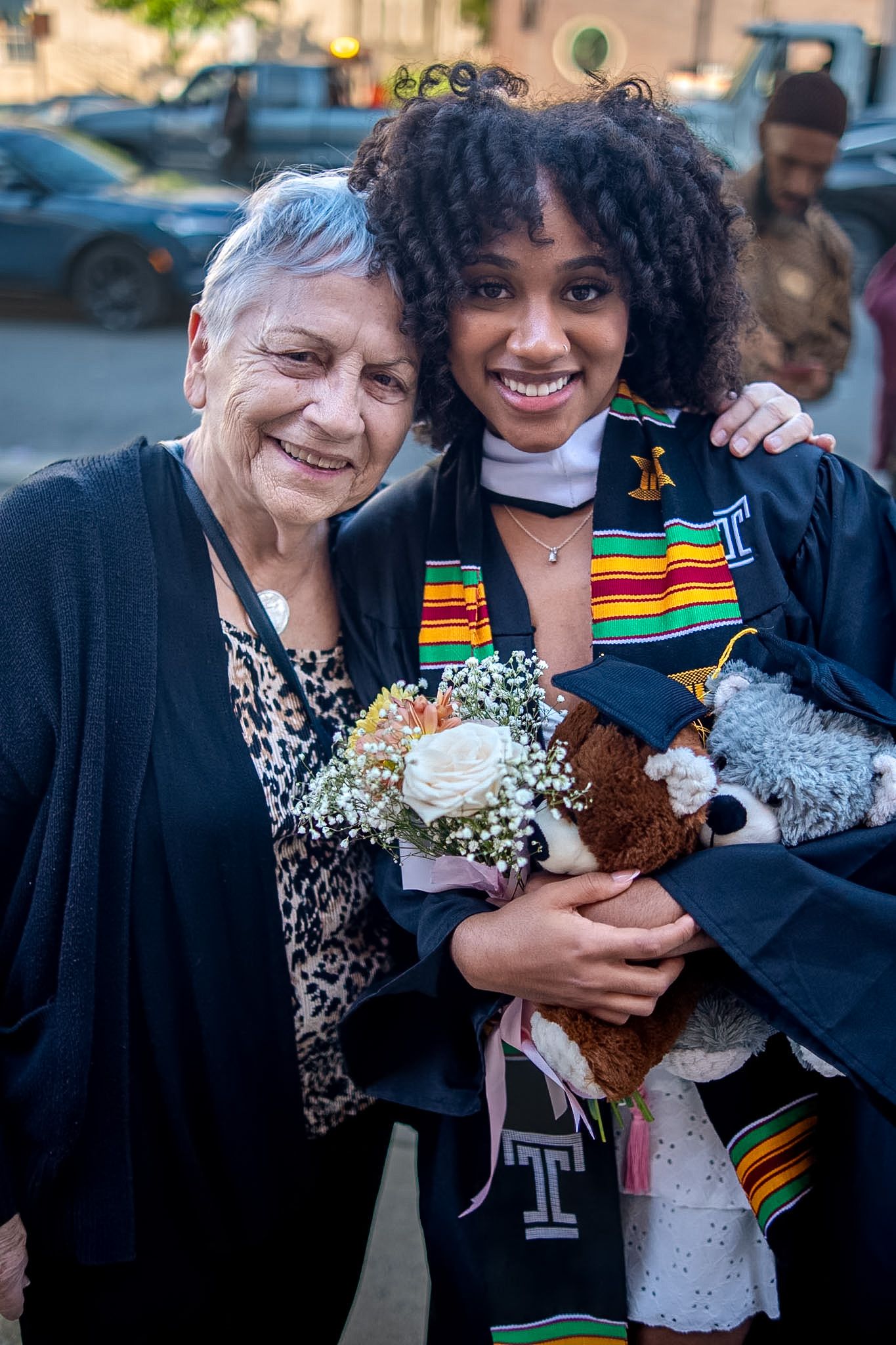 Elderly woman and young graduate in cap and gown holding flowers and teddy bear, smiling together at graduation ceremony.