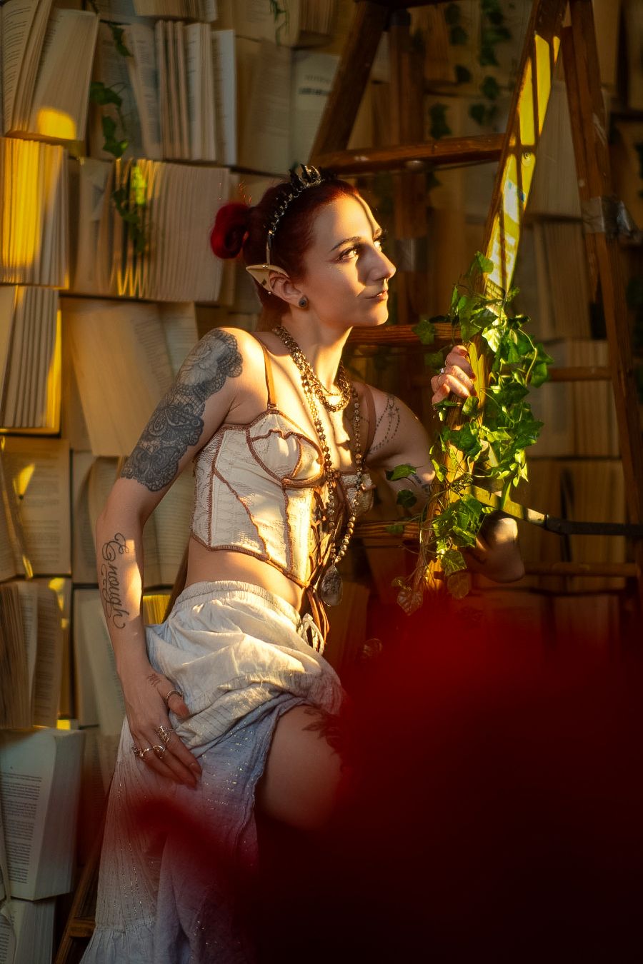 Fantasy elf cosplay woman with pointed ears, tattoos, and ivy in a warm library setting with stacked books.