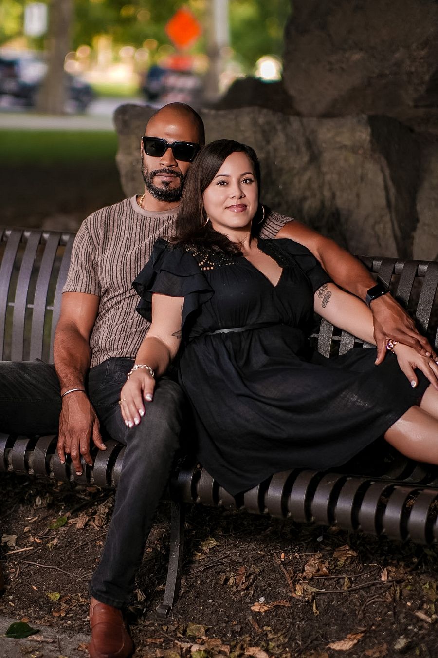A couple sits closely on a park bench, man in sunglasses and striped shirt, woman in black dress, sharing a tender moment.
