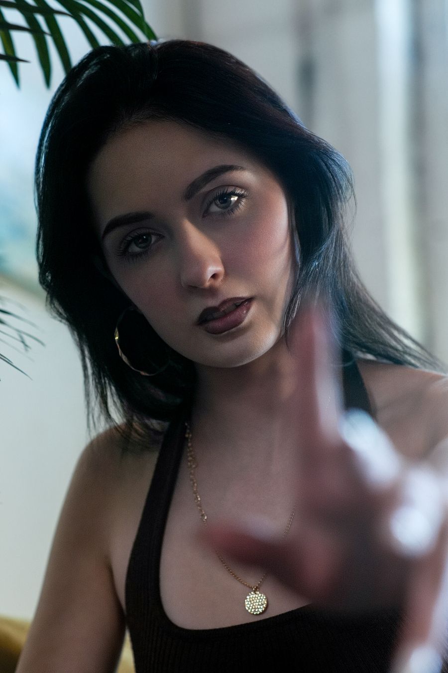 Dark-haired woman in brown tank top with gold necklace reaches toward camera in moody portrait.