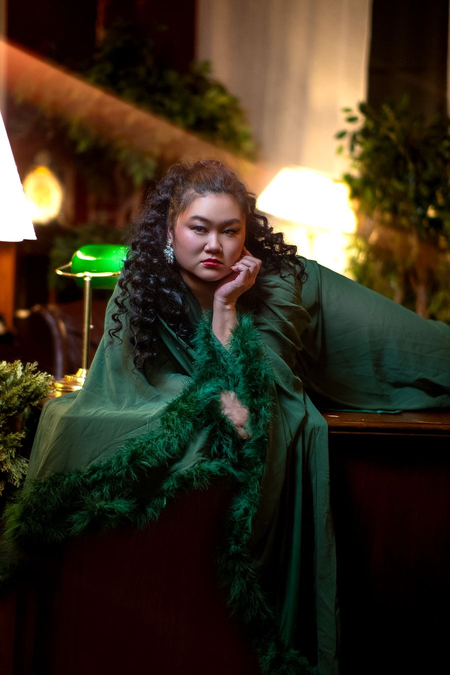 Woman in elegant green gown with feather boa, posing thoughtfully in a warmly lit, plant-filled interior setting.