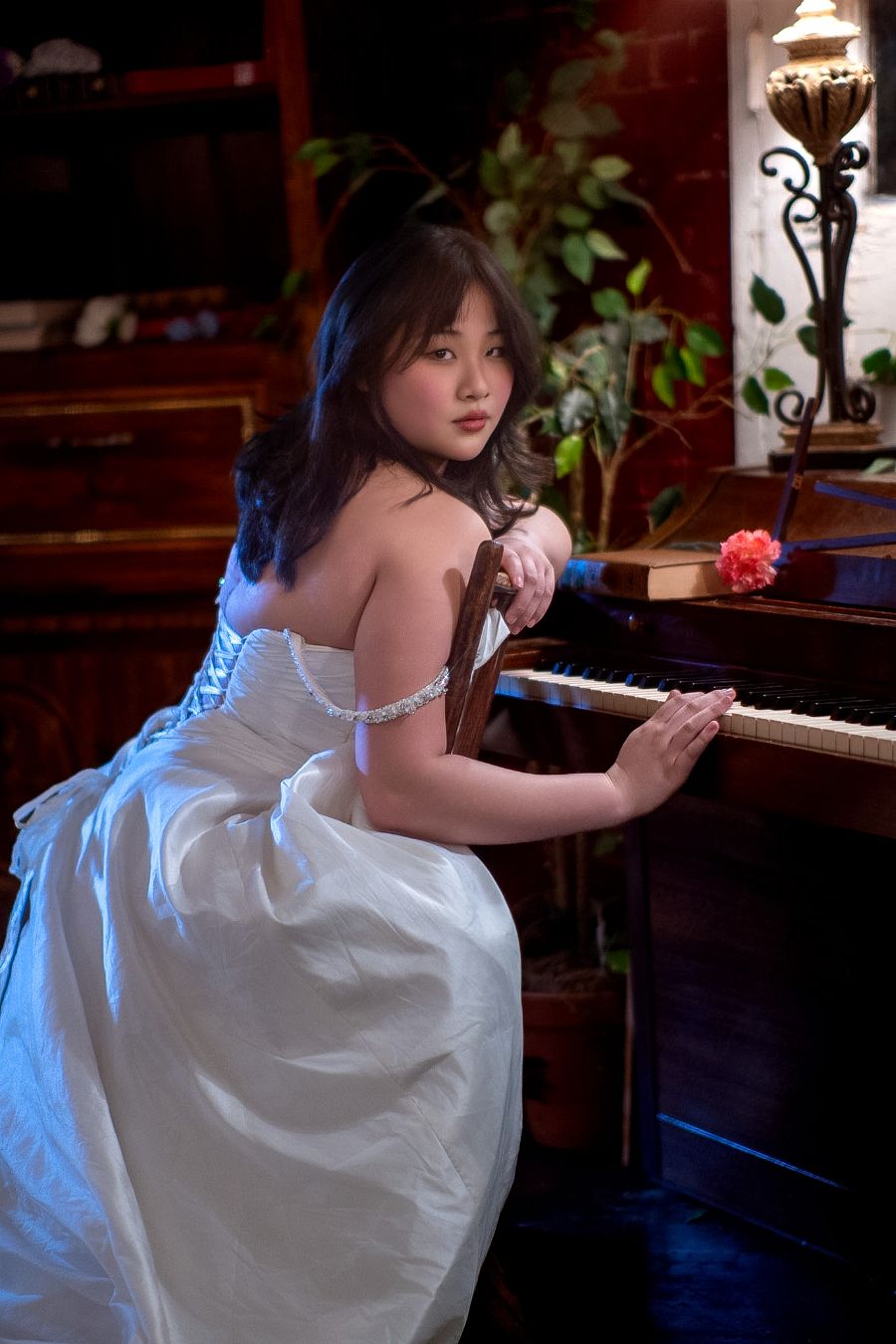 Young Asian woman in white strapless ball gown seated at a grand piano, looking over her shoulder elegantly.