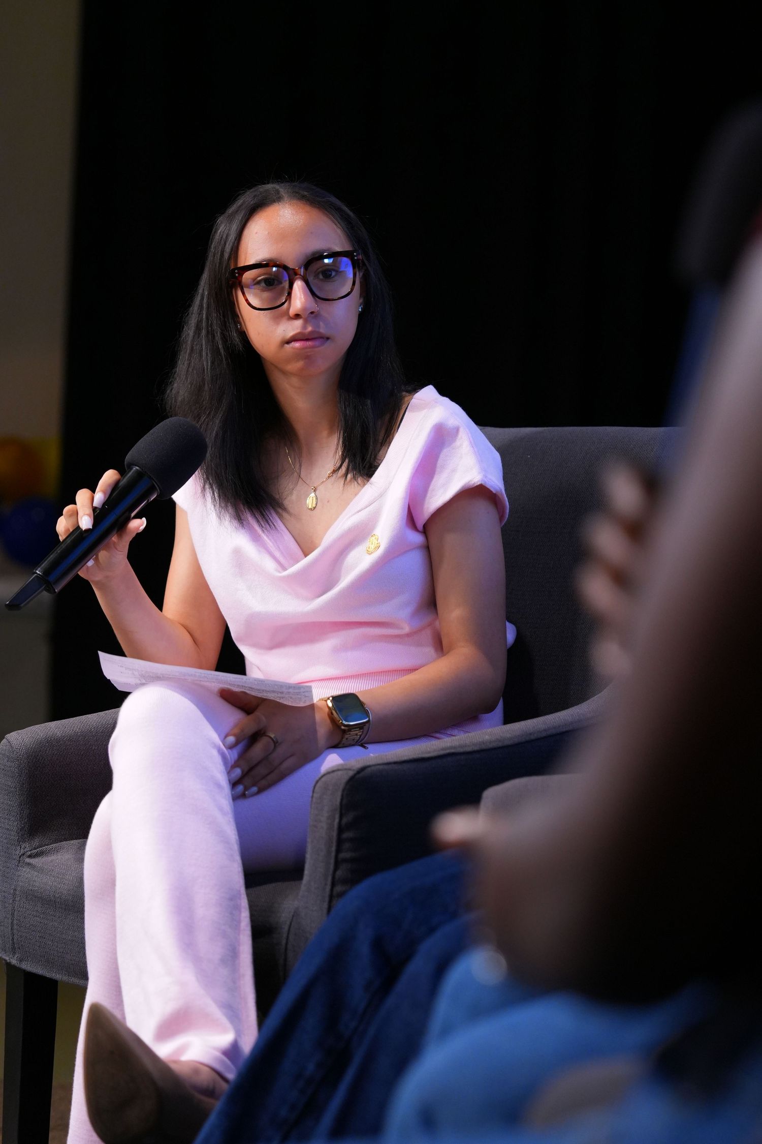 Young woman in pink outfit holding microphone while seated in gray chair during panel discussion event.