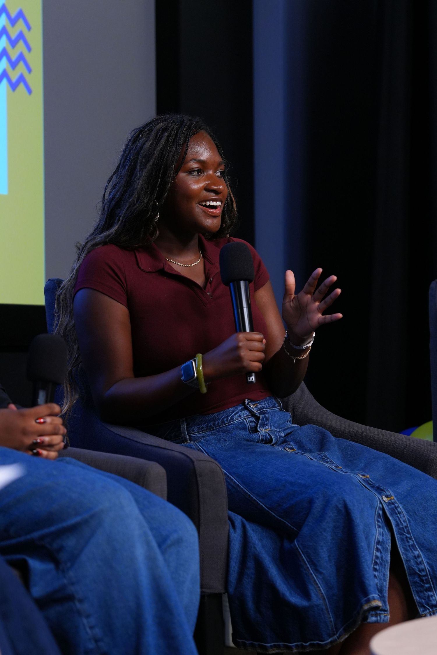 Young woman speaking into a microphone at a panel discussion, wearing a maroon top and denim skirt.