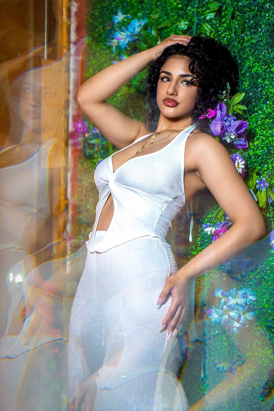 Woman in white crop top and skirt poses against floral wall backdrop with colorful flowers.