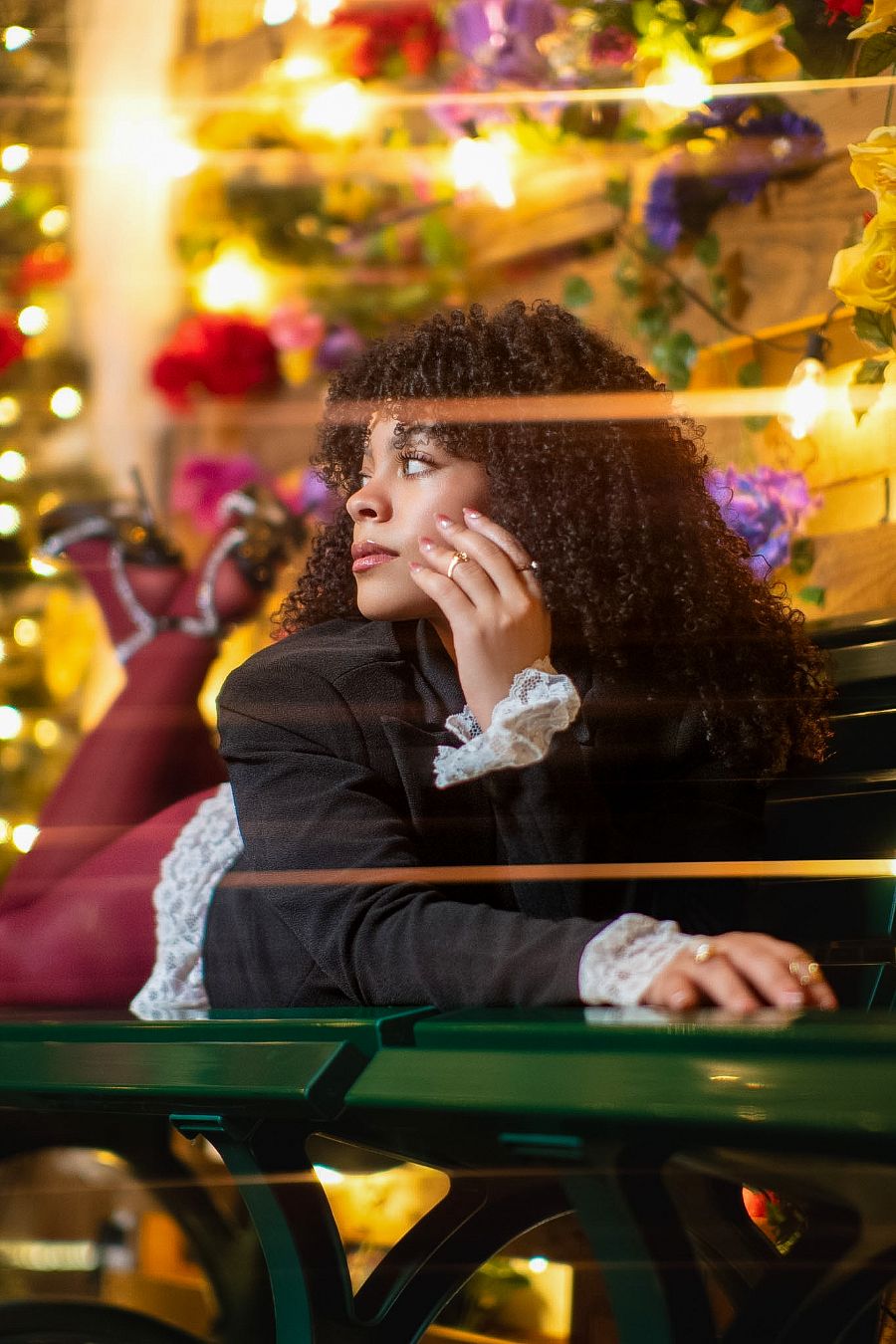 Young Black woman with curly hair sits pensively at a cafe table, surrounded by colorful flowers and warm string lights.