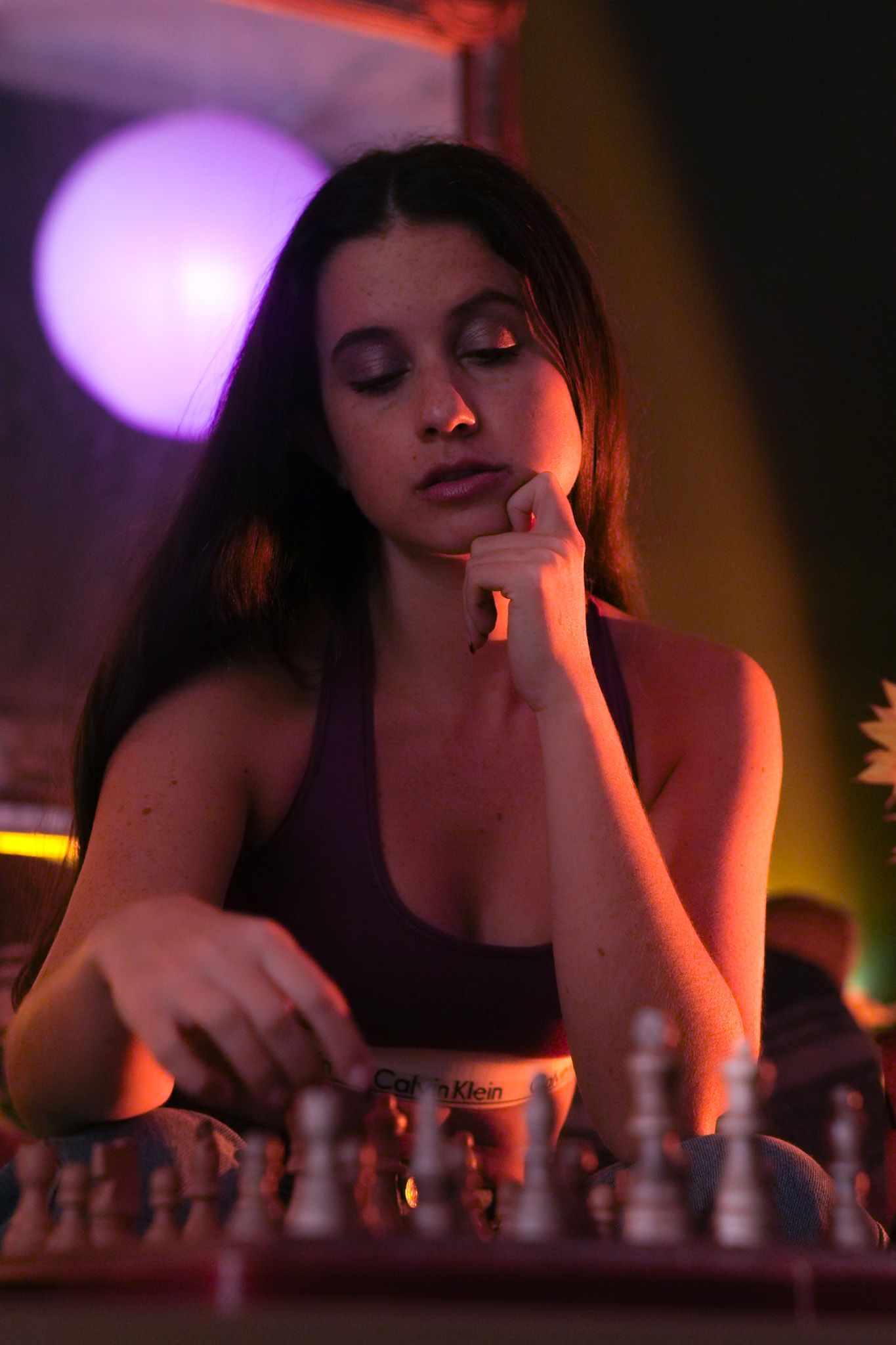 Young woman playing chess in moody purple and orange neon lighting, resting chin on hand in deep concentration.