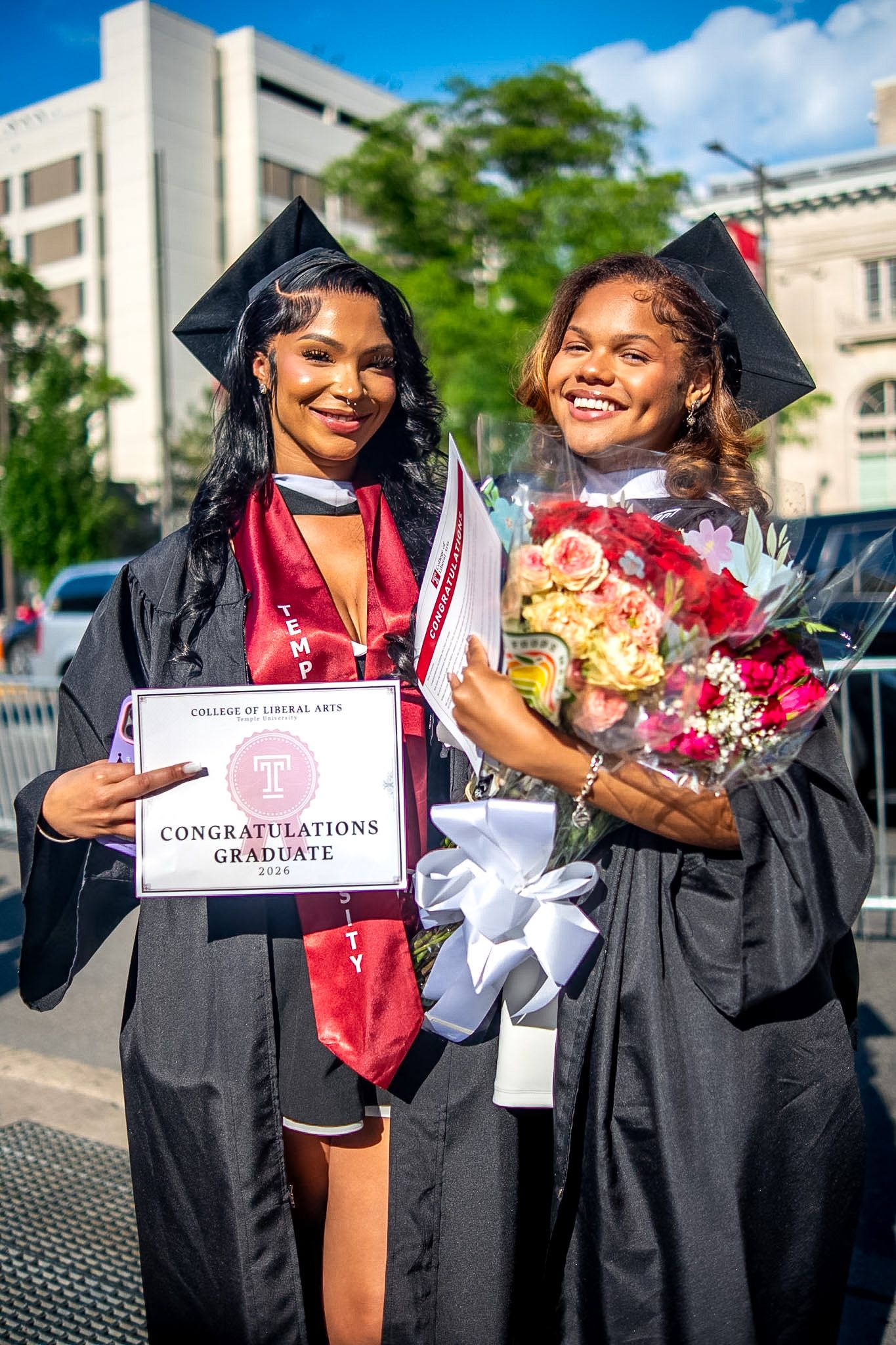 Two Temple University graduates in caps and gowns smile holding flowers and a Congratulations Graduate sign.
