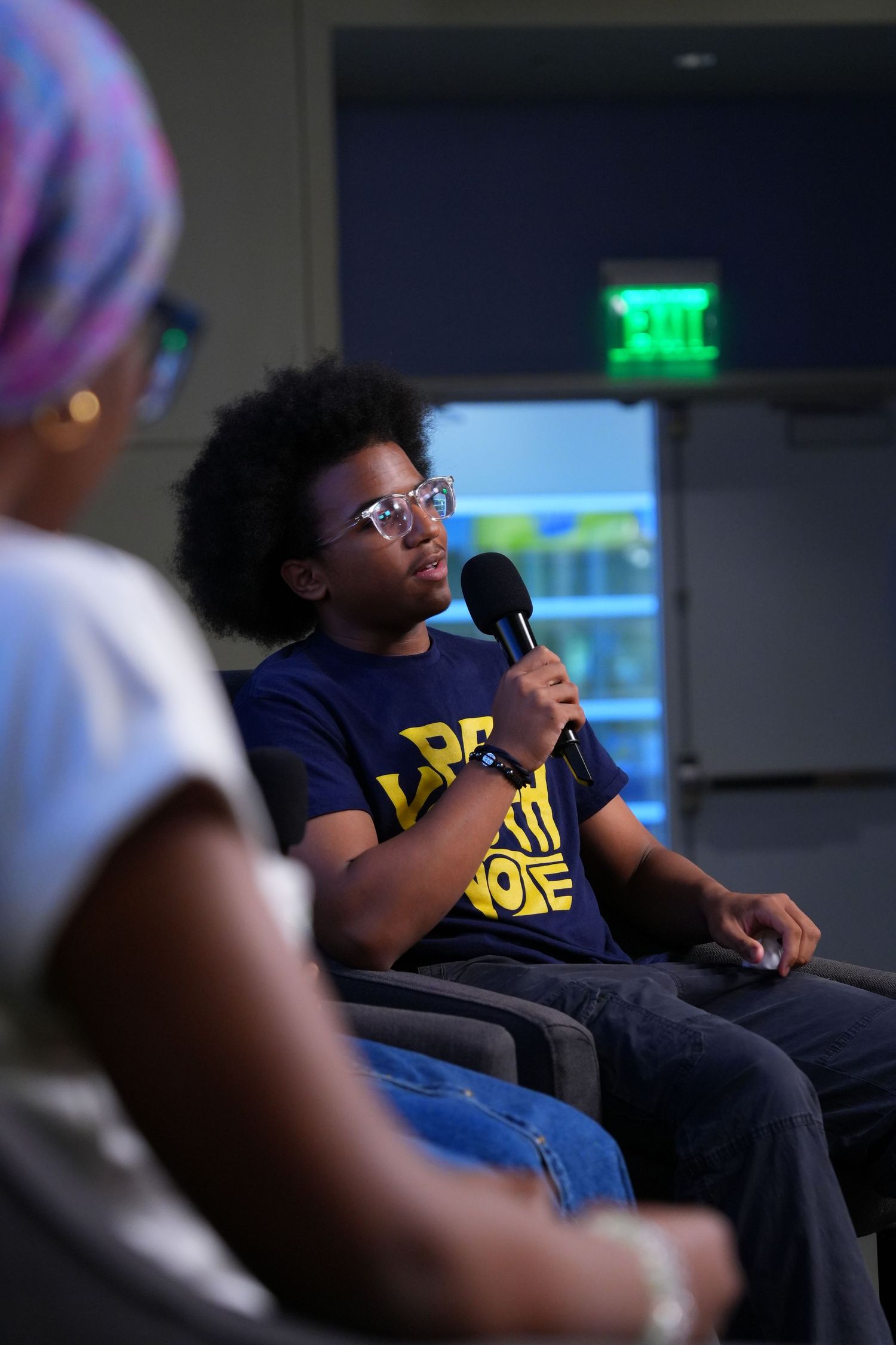 Young man with afro and glasses speaks into microphone at panel discussion, wearing a navy graphic tee.