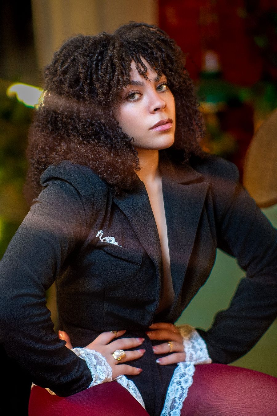 A stylish woman with curly hair poses confidently in a black blazer with lace accents and rings.