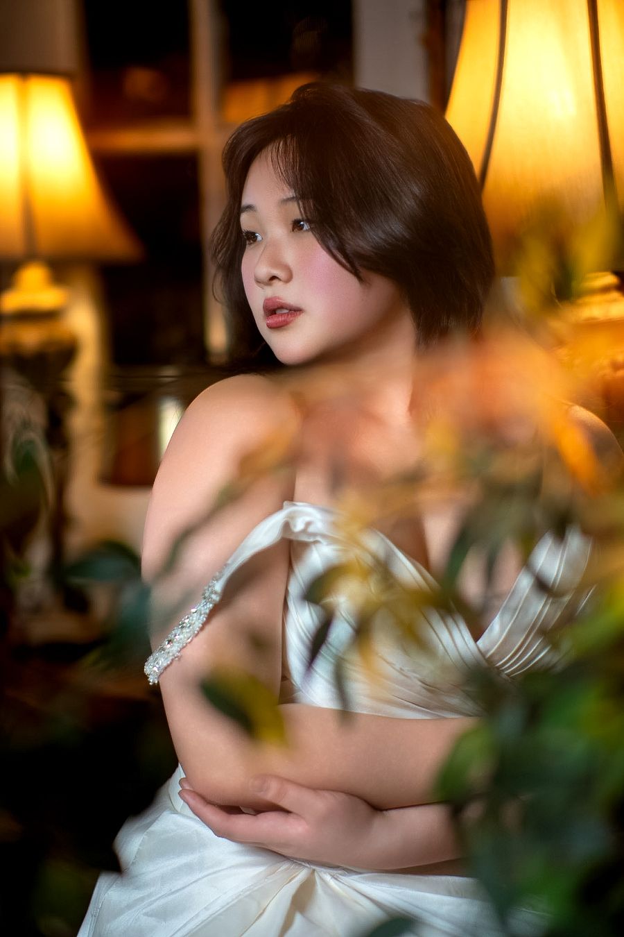 Young Asian woman in white satin dress sits thoughtfully among plants, bathed in warm lamp light indoors.