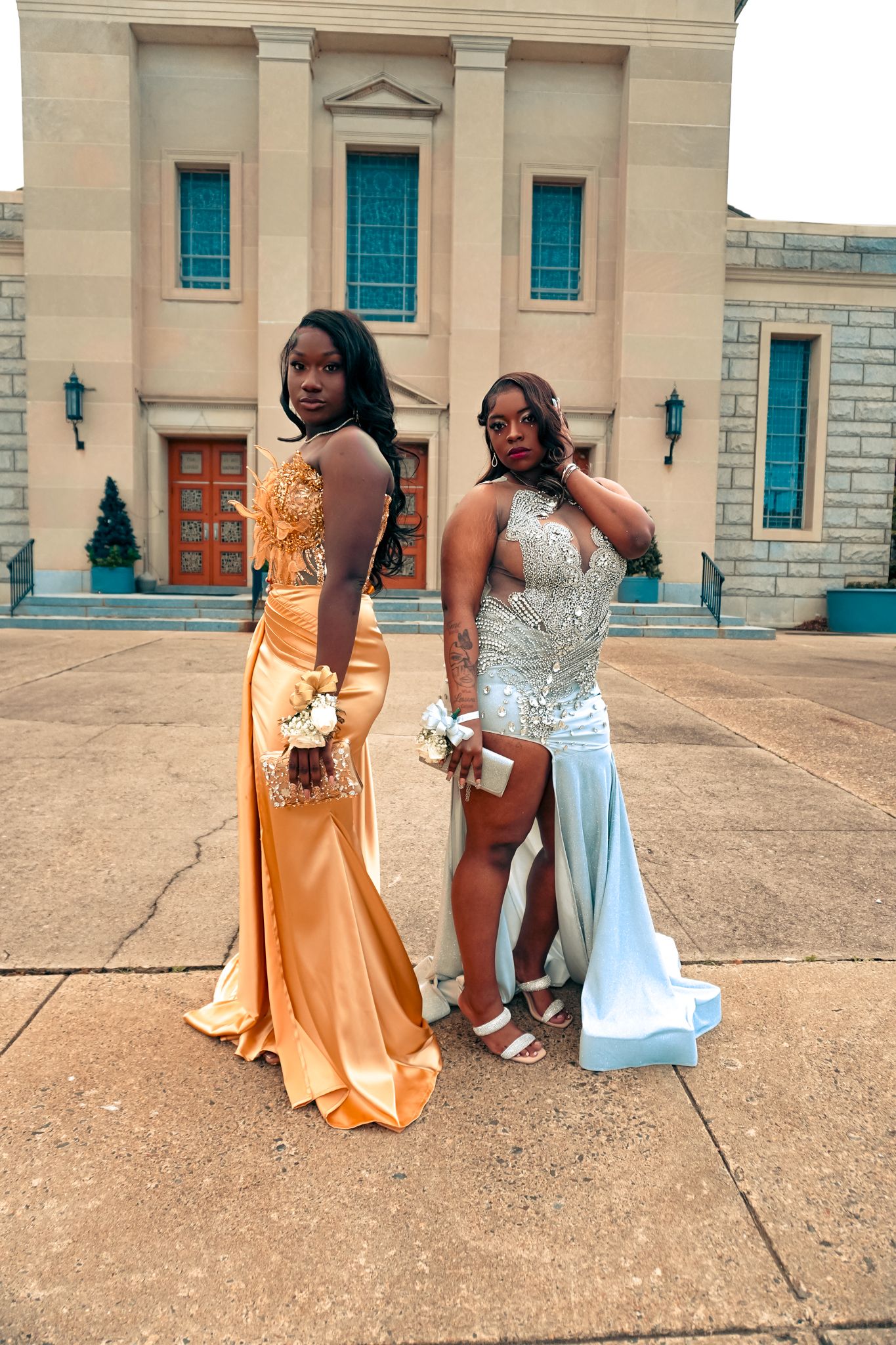 Two young Black women in elegant prom gowns, one gold and one silver, pose in front of a grand building.