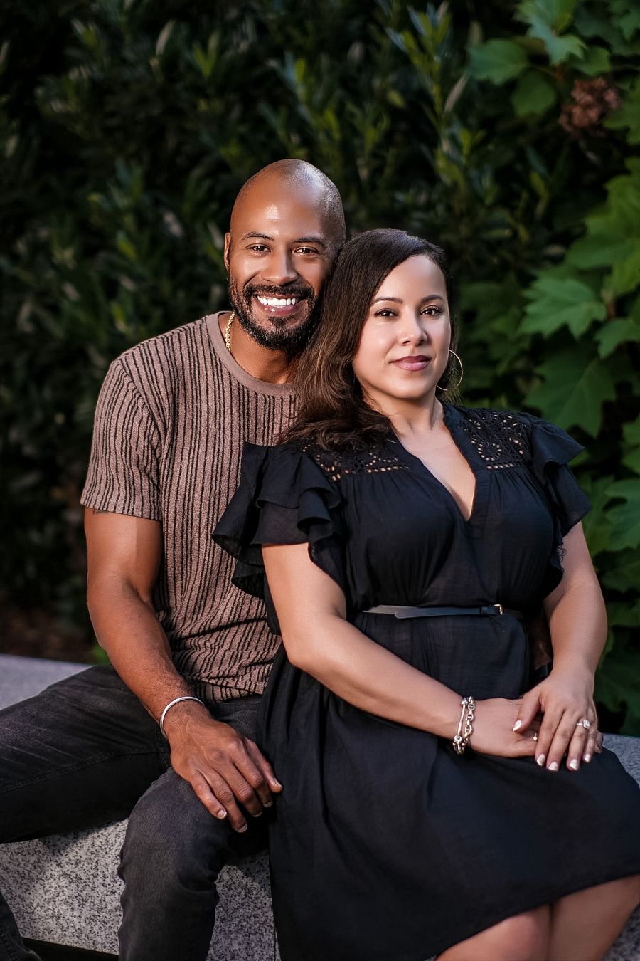 Smiling couple sitting together outdoors, man in brown shirt and woman in black dress, with green foliage background.