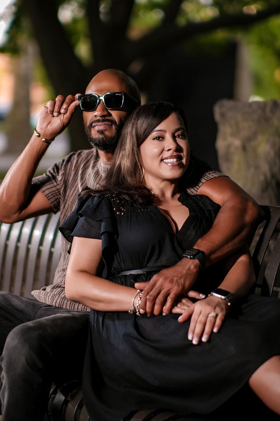 A couple sits on a bench outdoors, man in sunglasses with beard, woman in black dress, smiling together.