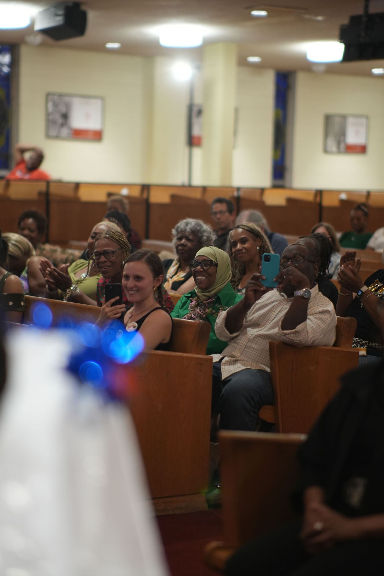 Congregation seated in church pews, many holding phones, during a lively religious service or ceremony.