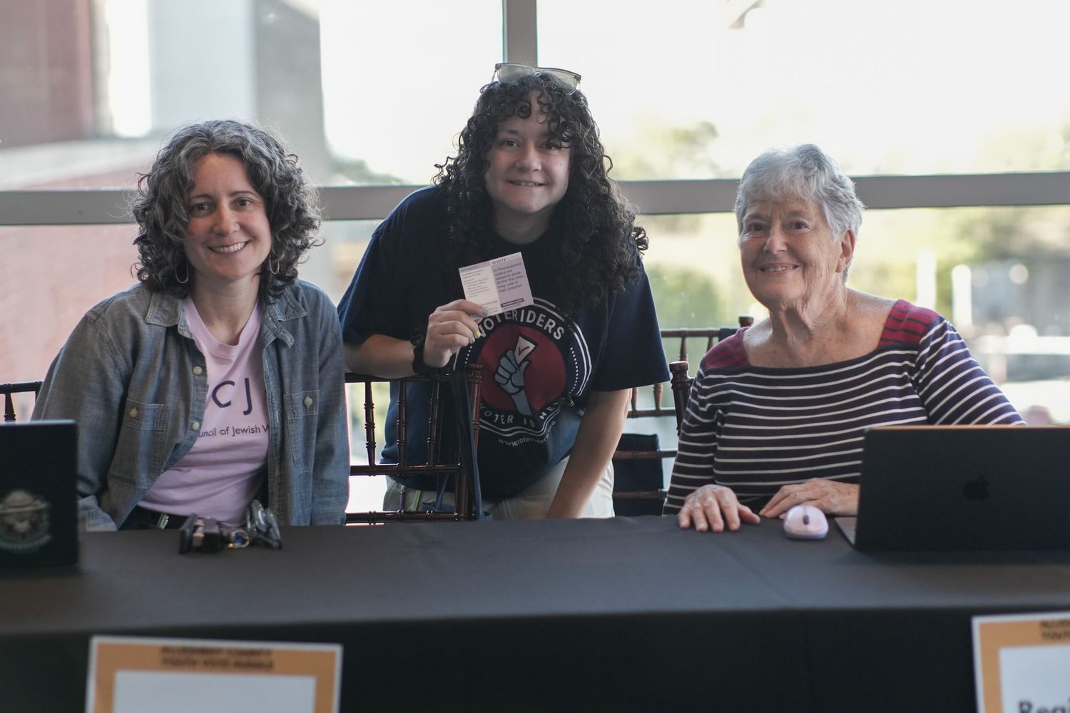 Three women smiling behind a registration table at an indoor event.