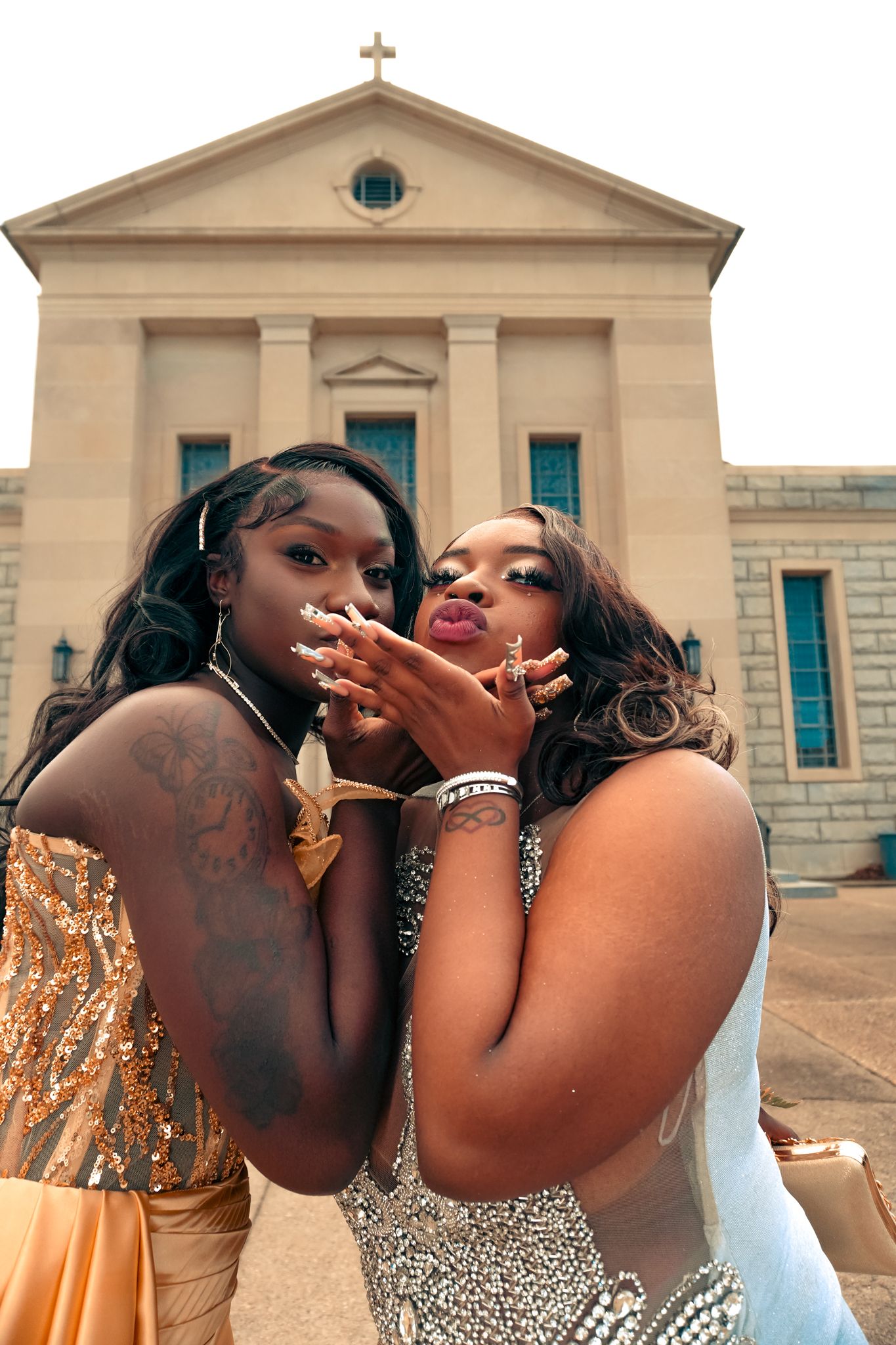 Two young Black women in sparkling prom dresses blow kisses toward the camera in front of a church building.