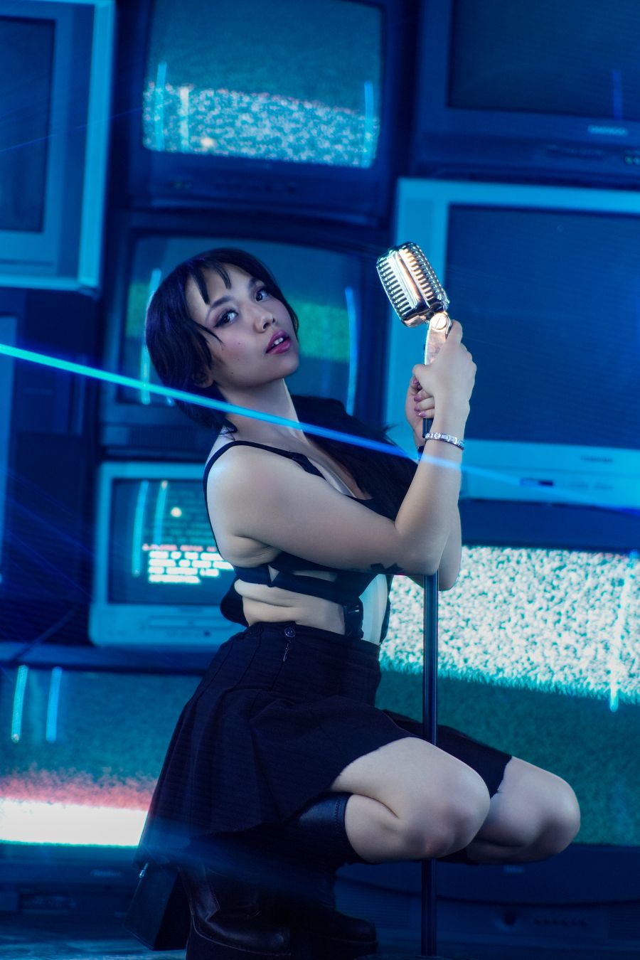 Young woman in black outfit holds vintage microphone on stand surrounded by retro TVs in blue light.