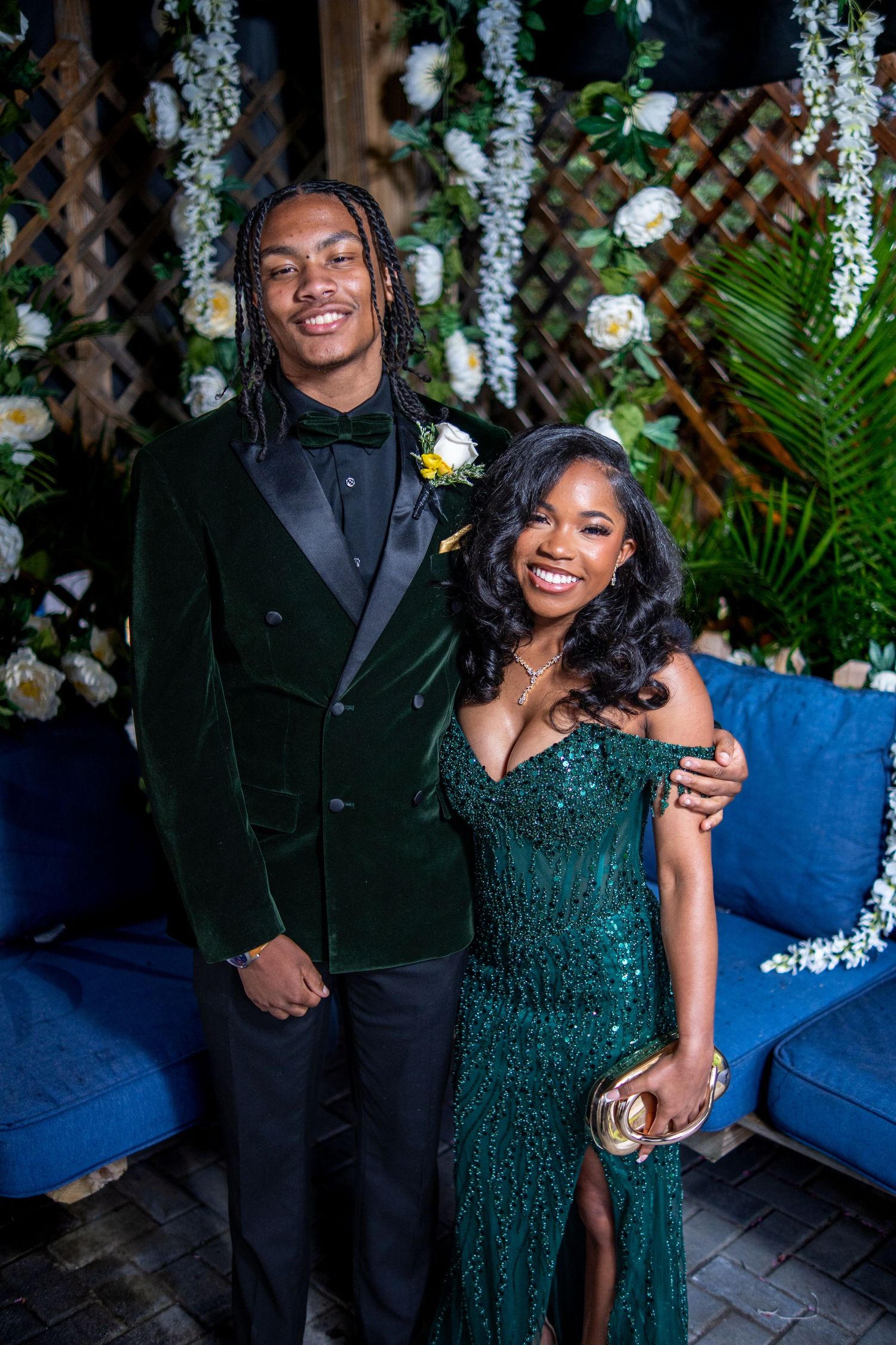 Couple in matching emerald green formal wear posing together at prom in front of floral backdrop.