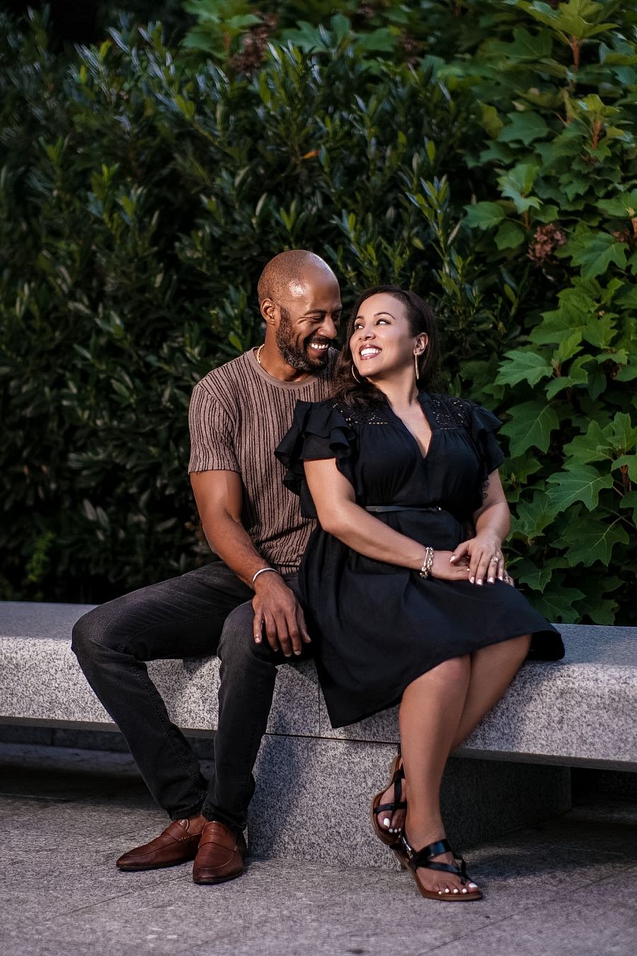 A smiling couple sits on a stone bench surrounded by lush greenery, sharing a joyful and intimate moment together.