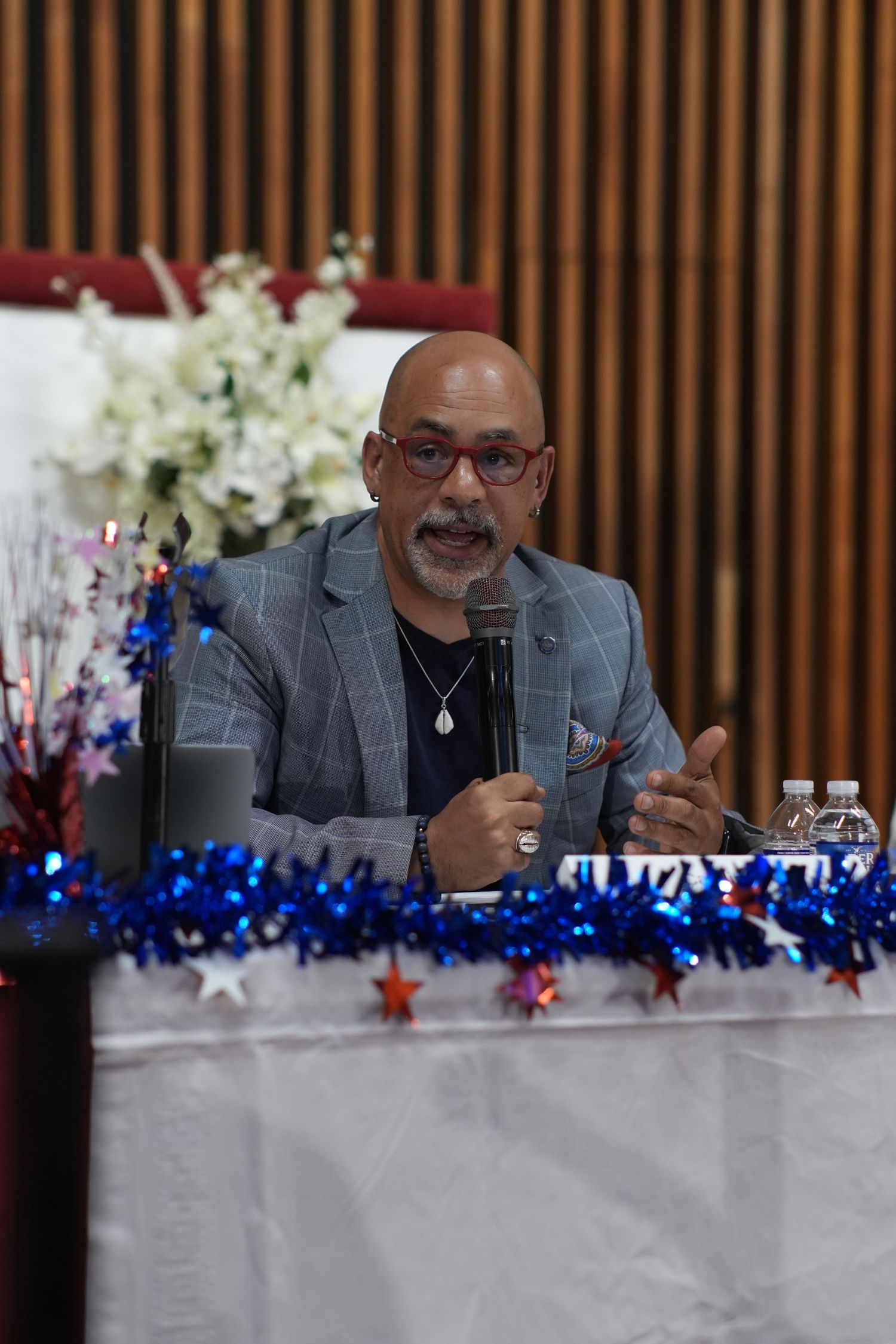 Man in gray blazer speaks into microphone at decorated table with patriotic blue tinsel and star decorations.