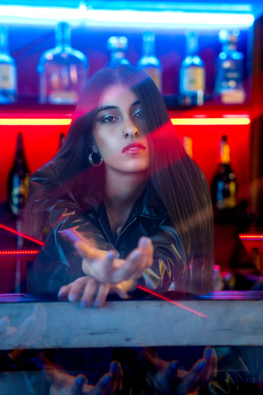 Woman in black leather jacket poses at neon-lit bar with red and blue lights and liquor bottles in background.