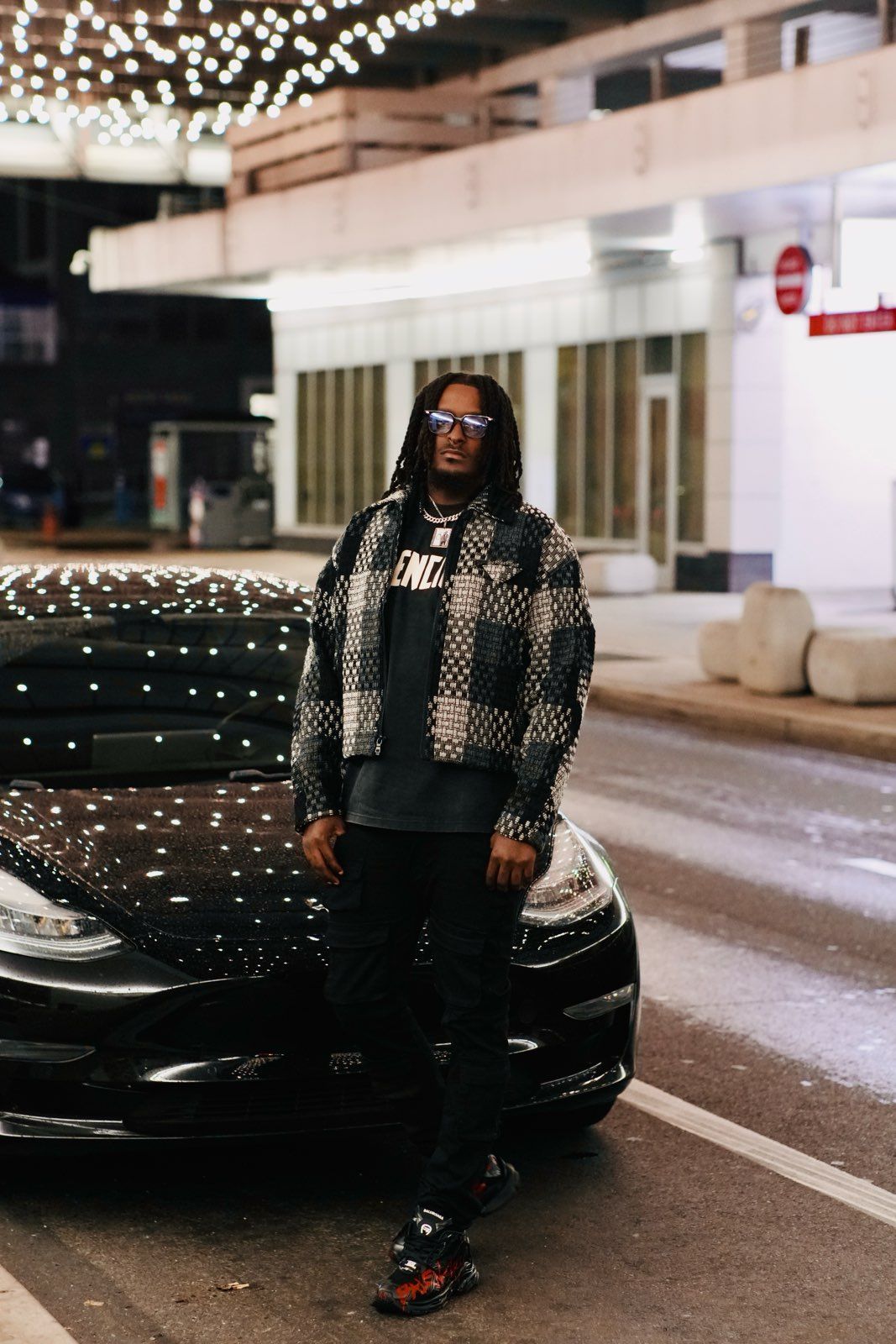 Stylish man in patterned jacket and blue sunglasses posing against black Tesla at night.